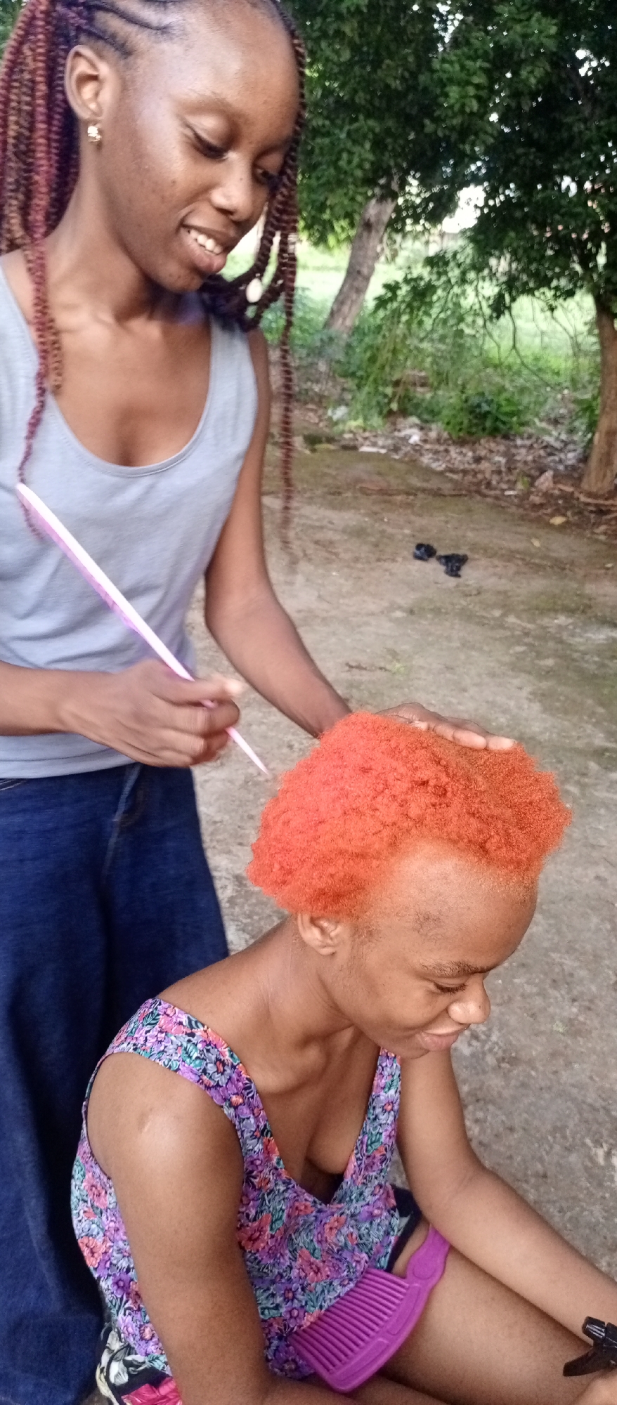 Two people are in an outdoor setting. One person is sitting, and the other is standing. The sitting person has short, orange hair, a floral top, and is looking down. The standing person has long, braided hair, a gray tank top, blue pants and is combing the sitting person's hair. The background has a blurred tree. The city location is Unknown, Unknown.