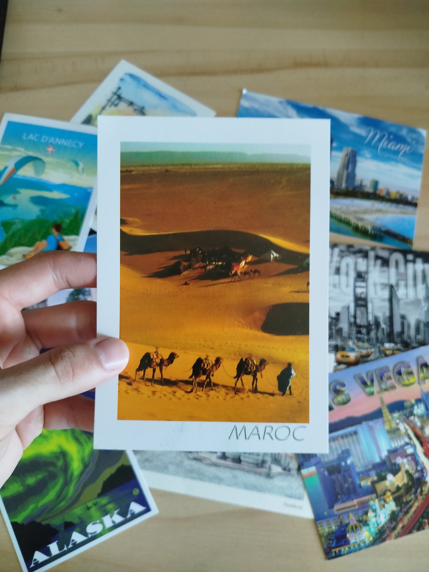 The image displays a collection of postcards arranged on a wooden surface. The most prominent postcard, held by a hand, features a desert landscape with several camels walking across sand dunes. The text "MAROC" is visible at the bottom of this postcard. Other visible postcards include one with "LAC D'ANNECY" and paragliders, another with "Miami" and city skyline, one with "New York City" and skyscrapers, and a postcard of "Alaska" with the Aurora Borealis. There is no indication of Varna, Bulgaria in the image.
