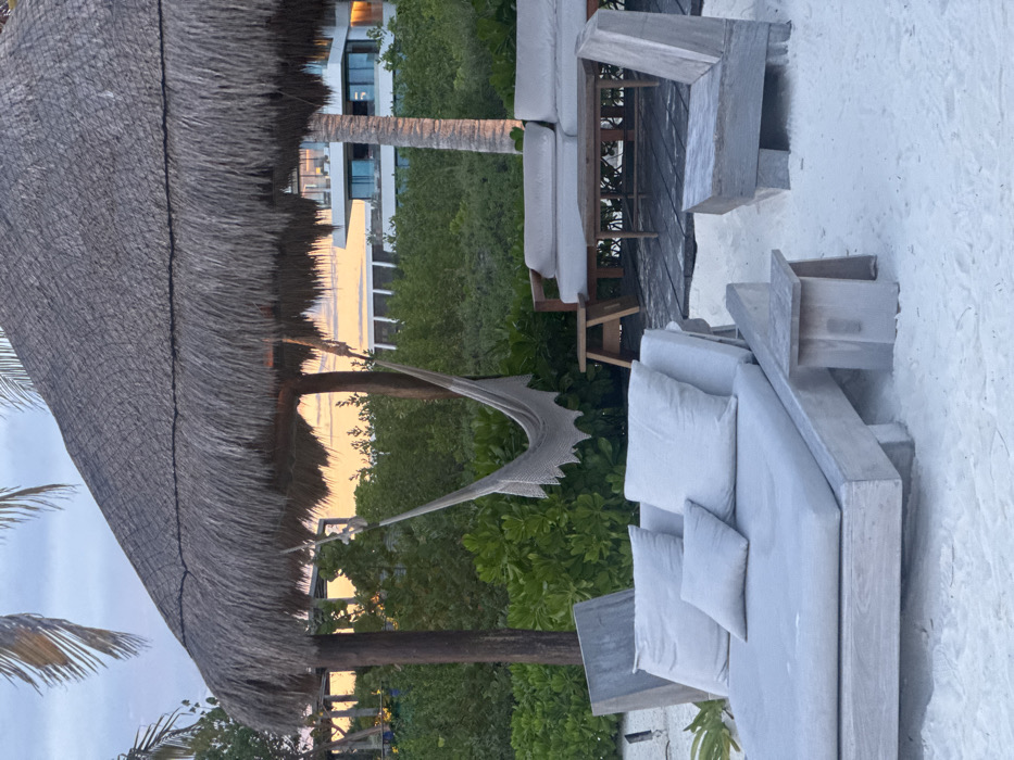 A beach scene shows white sand and pale gray outdoor furniture including sofas and lounge chairs. A thatched roof structure provides shade. Lush green foliage and trees fill the background, with a tall palm tree and a multi-story building visible further back. A white hammock hangs suspended. The sky appears overcast with hints of orange light, possibly indicating sunrise or sunset.