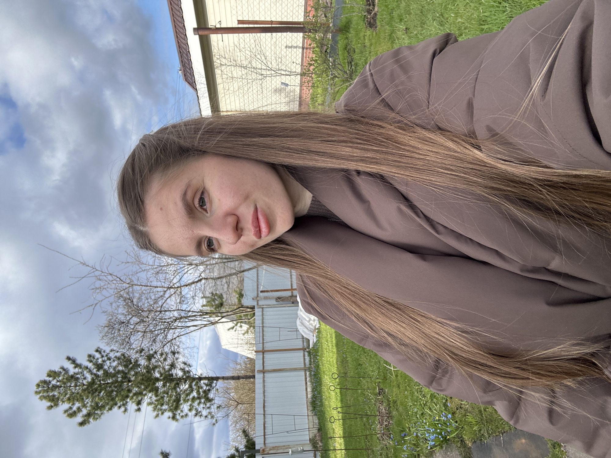 A portrait-oriented selfie captures a young woman outdoors in Klin, Russia, under a partially cloudy sky. The woman, positioned in the foreground and center-left, is seen from the chest up, gazing directly into the camera with a neutral expression and a slight pout. She has long, straight, light brown hair that flows over her shoulders and a dark brown or charcoal gray puffer jacket. Her eyes are light brown or hazel.
The background reveals an outdoor setting, likely a backyard or garden. To her left, behind her, and extending to the lower right, is green grass interspersed with small blue spring flowers, possibly forget-me-nots. Several thin metal garden stakes with circular tops are visible in a bed of dirt and sparse grass. Further back, a fence constructed from both chain-link and solid panels runs horizontally, separating the immediate area from a light-colored building, which could be a house or an outbuilding. Beyond the fence, several bare deciduous trees and a small evergreen pine tree indicate early spring conditions. The sky is a mix of white clouds and patches of blue, suggesting daytime. No text is visible in the image.