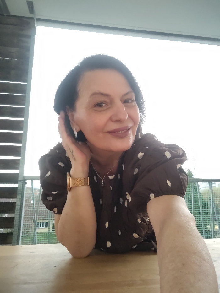 A woman smiles warmly, resting her cheek on her hand. She wears a brown, polka-dotted dress, a rose gold watch, and a delicate silver necklace. The background is a bright, overcast day with hints of green trees and buildings visible through a balcony railing. She stands beside a wooden table, and is framed by a wooden latticework.