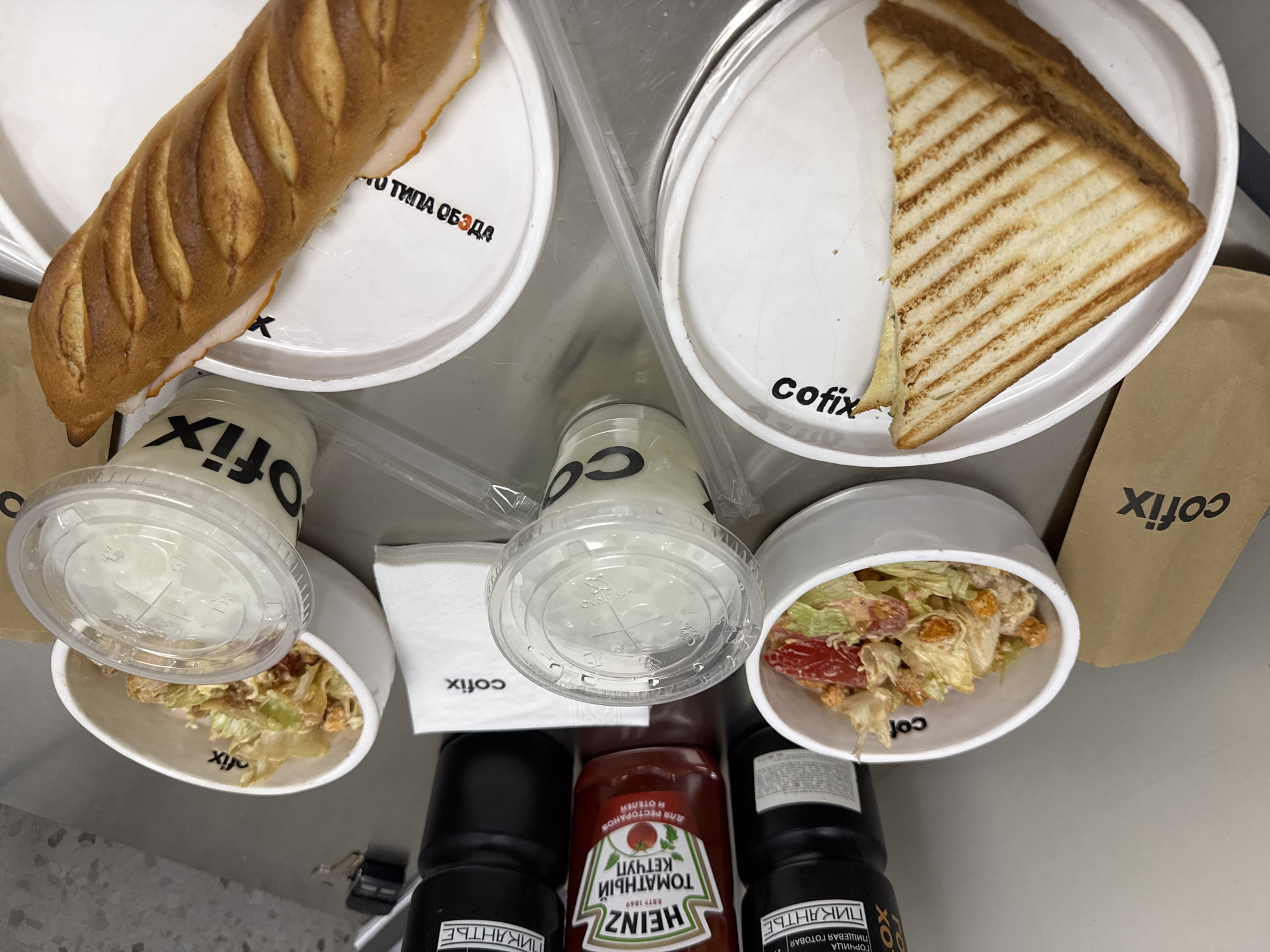 The image shows a fast-food meal on a table. The meal appears to be from the chain "cofix," judging from the branding on various items. The food consists of a baguette sandwich, a toasted sandwich, a salad in a bowl with a lid, and two drinks in cups with lids. Condiments like Heinz ketchup and prepared mustard ("Пикантье") are visible. The presence of Russian text on the mustard and on the plate with the baguette sandwich suggests that the location is not the United States or another primary English-speaking country. The presence of "cofix" branding suggests that the location may be in Yerevan, Armenia, as this chain has locations there. The scene is indoors, possibly in a restaurant or food court. No people are visible in the image.