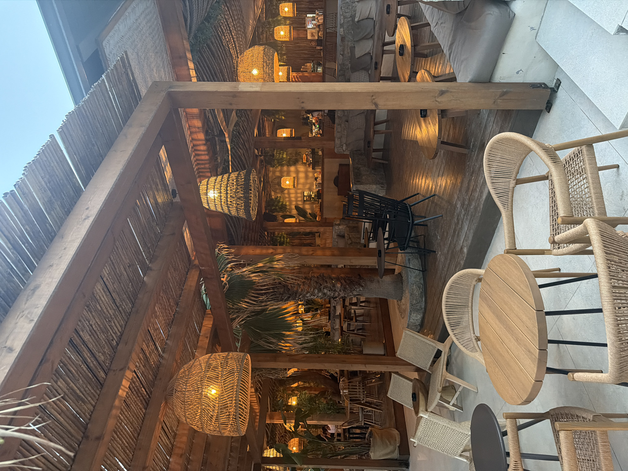 The image depicts an outdoor seating area of a restaurant or bar, likely located in Sithonia Municipal Unit, Greece, given the architectural style and vegetation. The scene is captured at dusk or early evening, with warm artificial lighting illuminating the space.
The primary focus is on the seating arrangements, which include several wooden tables and chairs. In the foreground, a round wooden table with a slatted top is surrounded by two woven chairs. To the left, a dark circular table with a metal base is partially visible, along with more woven chairs and a reclined woven chair.
The seating area is covered by a rustic wooden pergola with a thatched roof, creating a natural and tropical ambiance. Large woven pendant lamps hang from the pergola, casting a warm glow. Tropical plants, including palm trees, are strategically placed throughout the space, enhancing the outdoor feel.
In the background, a bar area is visible, with patrons seated on stools and staff attending to them. The walls are constructed from natural stone, adding to the rustic aesthetic. The floor is a combination of wooden decking and light-colored paving stones. The overall atmosphere is relaxed and inviting, suggesting a popular dining or lounge spot.