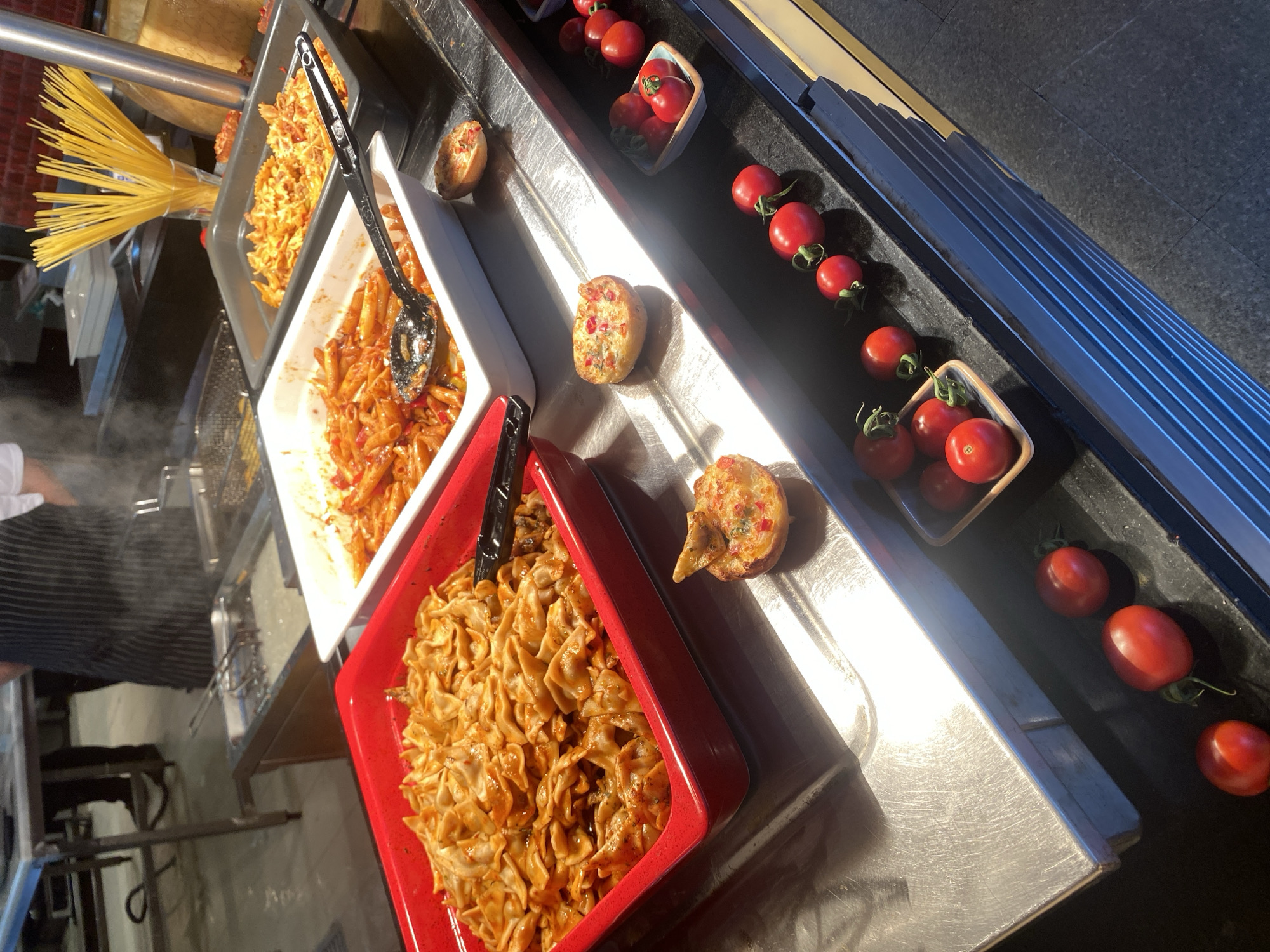 This is a close-up, eye-level shot of a buffet line in what appears to be a restaurant kitchen. The foreground is dominated by a stainless steel counter, with various food items displayed in chafing dishes and bowls.
In a bright red rectangular dish on the left, a generous portion of a pasta dish with a red sauce is piled high. The pasta appears to be a flat, folded noodle shape. A black serving utensil rests on the dish.
To the right of the red dish is a white rectangular chafing dish filled with penne pasta in a red sauce with visible pieces of red pepper. A black serving spoon is placed inside this dish. Behind this, a grey chafing dish contains another pasta dish, possibly with bow-tie pasta in a similar red sauce. In the background, a glass holder filled with uncooked spaghetti noodles stands upright.
Further to the right, a small, round pastry or bun topped with colorful diced ingredients, possibly vegetables and cheese, is visible on the counter. Below this, a small square bowl holds a cluster of bright red cherry tomatoes. A line of individual cherry tomatoes, still attached to their stems, extends across the counter, with more tomatoes in small decorative bowls.
In the background, out of focus, is a commercial kitchen environment. A person wearing a black and white striped apron is visible, standing near a stainless steel food preparation area with steam rising from it. Another person's legs and feet in dark shoes are visible on the left side.
The overall lighting suggests artificial light, with a warm glow illuminating the food and creating reflections on the stainless steel surfaces. The setting is clean and organized, typical of a food service establishment. The location is not specified within the image or its context, and therefore cannot be determined to be Aksu, Turkey.