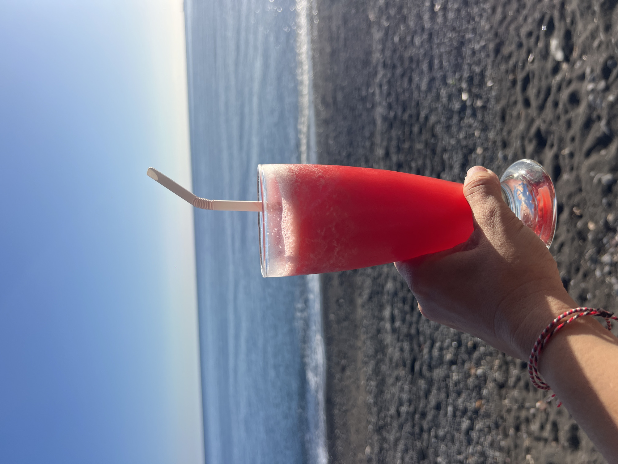 A close-up, outdoor, eye-level shot shows a hand holding a tall glass of bright pink, slushy drink with a pale pink straw. The drink is in focus in the foreground, with a blurred background of a beach and ocean. The beach is composed of dark pebbles and sand. The ocean is a deep blue with small waves and white foam at the shore. The sky above is a clear, bright blue. The hand has a thin, braided bracelet with red, white, and black threads. The scene appears to be on a sunny day. This image is not taken in the city of Abang, Indonesia.