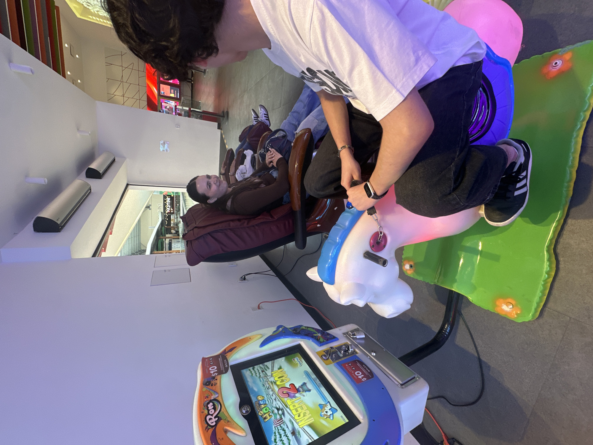 In a brightly lit indoor entertainment venue, likely an arcade or amusement center, in San Bernardino Tlaxcalancingo, Mexico, two young individuals are seated. To the left, a children's arcade game with a screen displaying a winter scene and text reading "INSERT 2 COIN" is positioned. Next to the game, a pink and white rocking horse-shaped ride is occupied by a person wearing a white t-shirt and black jeans. This person is holding onto handlebars and appears to be interacting with the game. In the background, another individual, a young woman with dark hair, is seated in a reclining massage chair, facing the arcade game and smiling. Other similar massage chairs are visible behind her, occupied by other patrons, suggesting a recreational environment. The floor is tiled with a dark gray pattern.