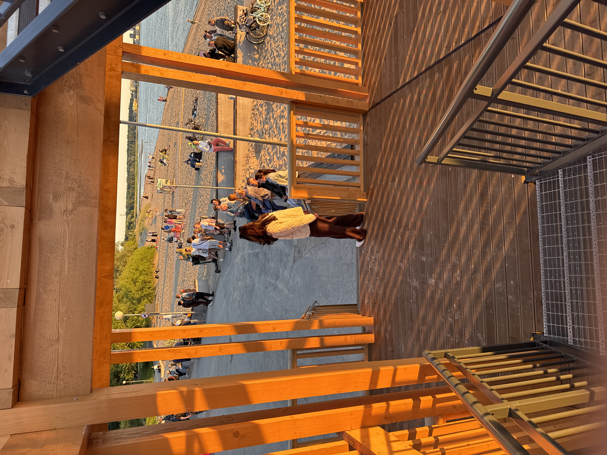 This image depicts a scene at a public beach or promenade area in Tallinn, Estonia, during sunset. The foreground shows a wooden deck with a metal railing and stairs, casting long shadows from wooden posts and railings due to the low angle of the sun. In the mid-ground, a woman in a light-colored puffer jacket and dark pants stands on the wooden deck, facing away from the camera. Beyond her, a sandy beach is populated with numerous people. Some are sitting on the sand or on benches, while others are standing or walking. A body of water, likely the Baltic Sea, is visible in the background, with a distant cityscape on the horizon. There are bicycles on the sand, and streetlights are also present. The overall atmosphere is leisurely, with people enjoying the evening light and the seaside environment.