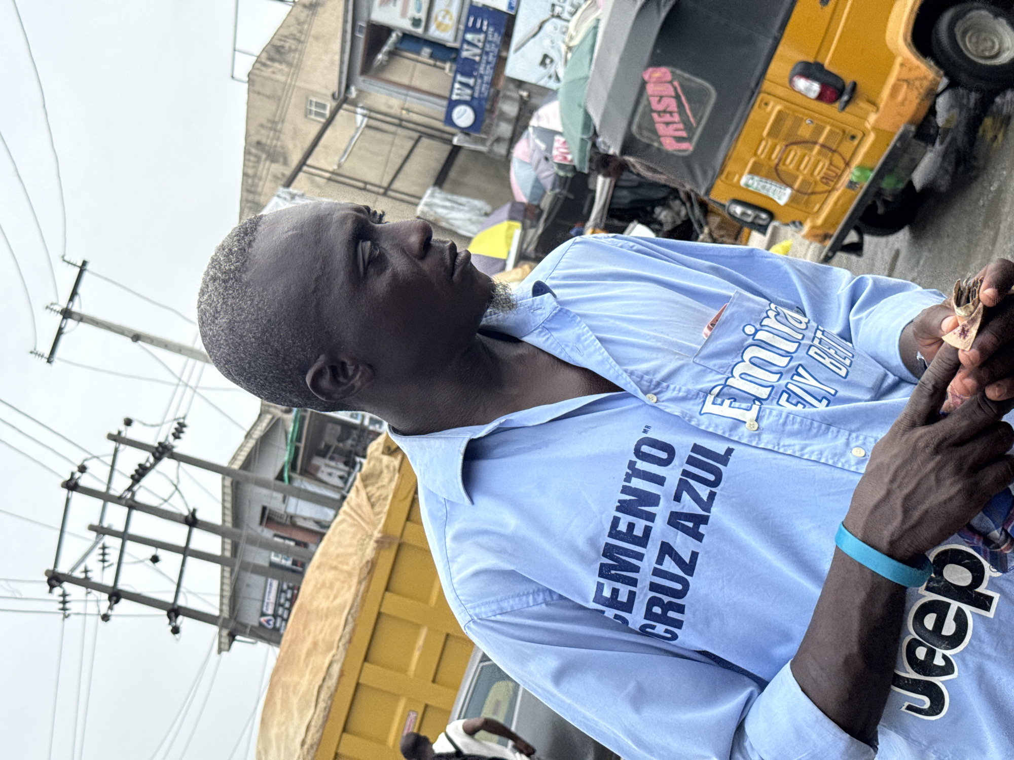 A medium shot shows a Black man looking upward to the right, holding an unidentified object in his right hand, standing on a street in Lagos, Nigeria. The man is wearing a light blue, button-down shirt with "CEMENTO CRUZ AZUL" printed vertically in large, dark blue letters on the left side. On the right side of the shirt, "Emirates Fly Better" is printed, and he is wearing a blue wristband. The man has short, graying hair and a goatee. A yellow "keke", or auto rickshaw, is partially visible behind him, as well as a partially visible yellow truck. Overhead power lines and buildings can be seen in the background.