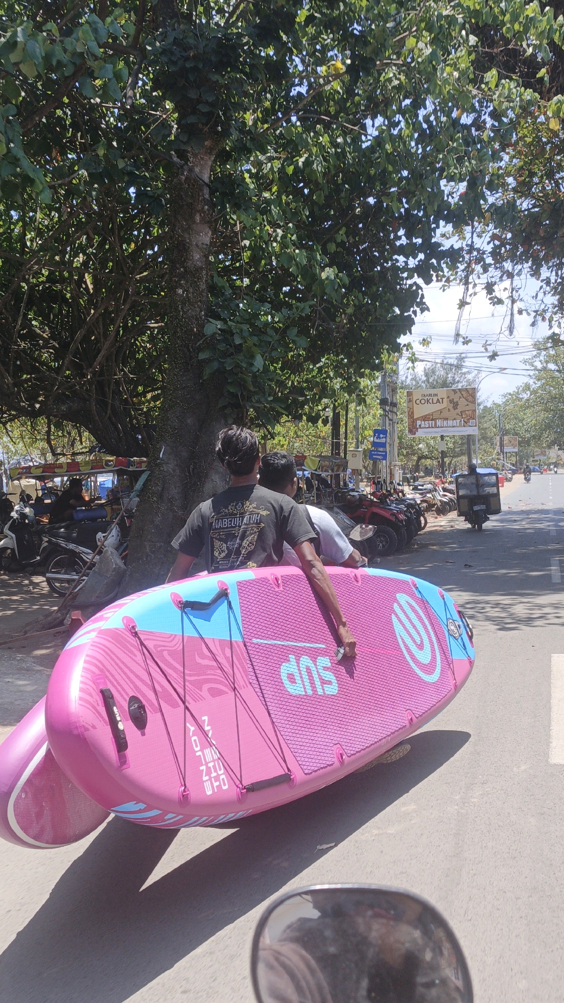 Two men are carrying a large, pink stand-up paddleboard (SUP) down a street in Desa Pagandaran, Indonesia. The man in the foreground, facing away from the camera, wears a black t-shirt with yellow text. The SUP has blue accents and the word "SUP" printed in blue on its surface. Behind them, a line of parked motorcycles and some ATVs are visible, as well as market stalls and a billboard advertising "DIARUM COKLAT" (Djarum Chocolate). The street is paved, and there is a sidewalk with trees and foliage. The sky is bright and sunny. A motorcycle mirror is visible in the bottom right corner of the frame, reflecting the scene.