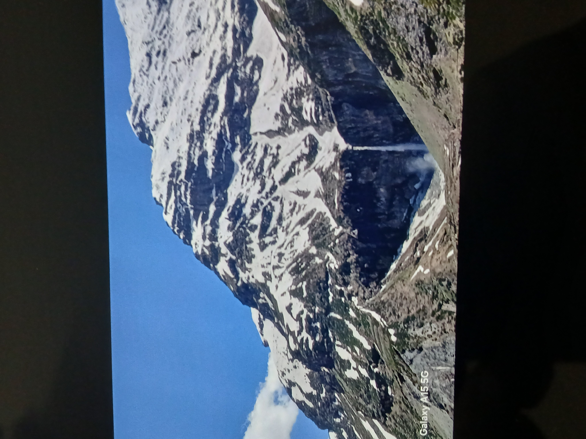 The image shows a tall, snow-covered mountain against a bright blue sky. The mountain appears rugged with visible rock formations and patches of snow. A waterfall or stream can be seen cascading down one side. The photo was taken using a Galaxy A15 5G phone. The location is not in Bengaluru, India, as the terrain is incompatible.
