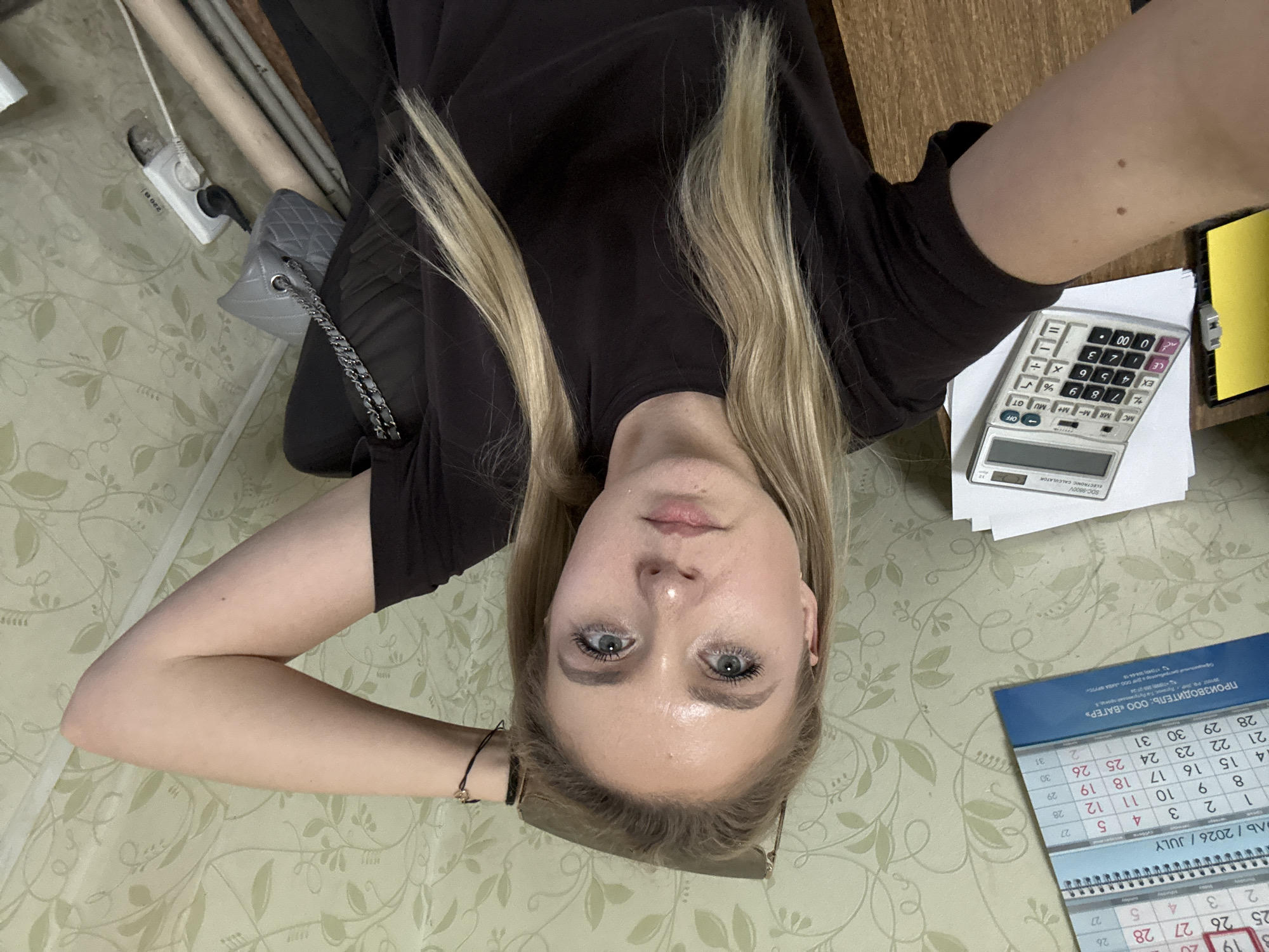 A woman is taking a selfie, holding the camera above her head. She has blonde hair and blue eyes, and appears to be wearing dark clothing. She is sitting in a chair with a quilted gray purse next to her. The background shows a wall with a floral wallpaper pattern and a power outlet with a plug. On the right side of the image, there is a wooden desk with a calculator and a calendar. The calendar indicates the month of July 2026. The lighting suggests it is daytime, and the overall scene appears to be indoors, possibly in a home or office environment. The woman's expression is neutral. Visible text includes "SOC-9800V ELECTRONIC CALCULATOR" on the calculator and "ПРОИЗВОДИТЕЛЬ: ООО «ВАГЕР»" and "ЛЬ / 2026 / JULY" on the calendar.