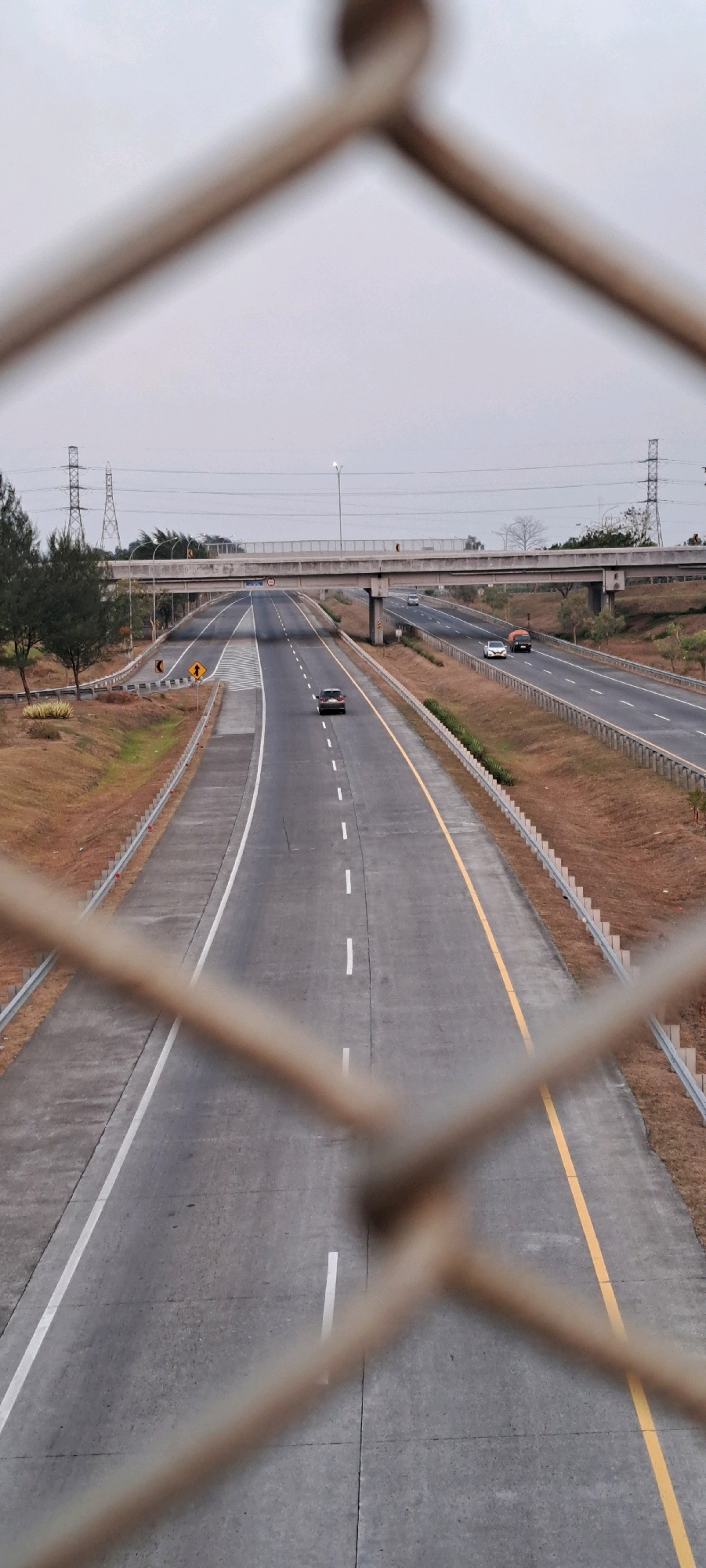 A high-angle, vertical shot captures a wide, empty highway in Sidowayah, Indonesia, with traffic moving in both directions. The road is bordered by a dry, brown grassy median and shoulder, with a metal fence in the foreground partially obscuring the view. A concrete overpass crosses the highway in the distance, with electrical towers and sparse trees dotting the landscape under a hazy, overcast sky. Several cars, a truck, and a dark-colored SUV are visible on the highway.