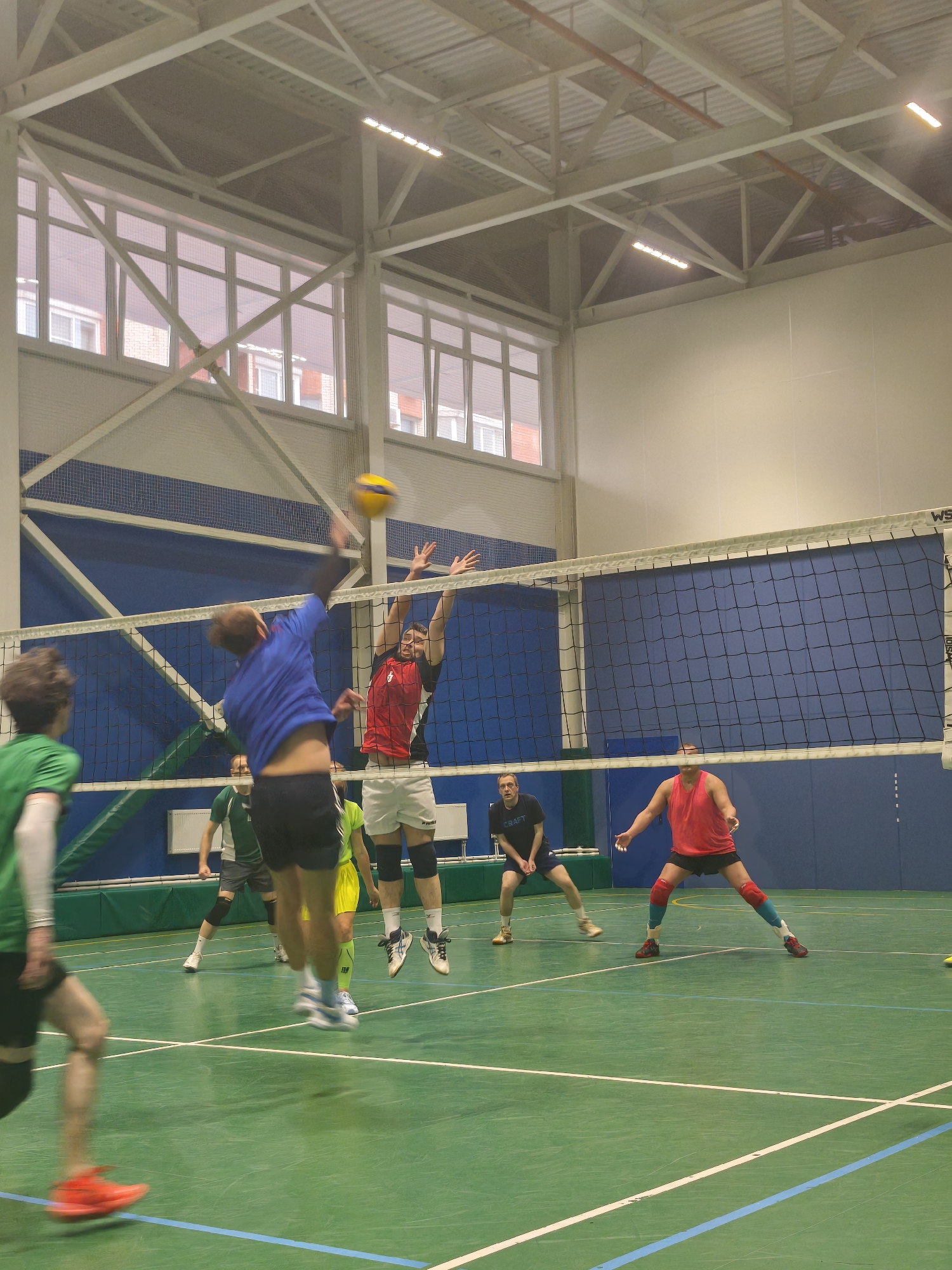The image depicts a volleyball game in progress inside a gymnasium. Several individuals are engaged in the game, with one player in a blue shirt spiking the ball over the net, while another player in a red shirt attempts to block. Other players are positioned on the court, ready to react to the play. The gymnasium has a green floor with blue lines, a volleyball net in the center, and large windows along the wall. The location could be in Saint Petersburg, Russia, based on architectural features that are common in Russia.