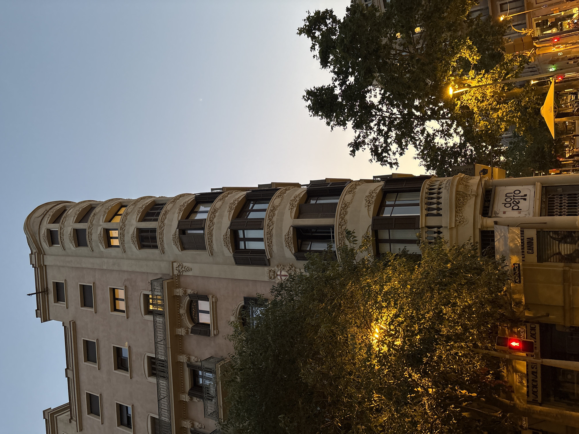 The image shows a corner building in Barcelona, Spain, with a distinctive curved facade. The building has multiple stories with windows, some of which are illuminated, suggesting it is either evening or early morning. The architecture features ornate detailing around the windows and balconies. In the foreground, there are trees with dense foliage, and a street lamp casts a warm yellow light on the leaves. A red traffic light is visible, showing a pedestrian signal. Signs are present on the lower parts of the building, including one that reads "don perro". The sky above is a gradient of pale blue and orange, indicating a twilight setting. The overall impression is of an urban street scene in Barcelona during either dawn or dusk.
