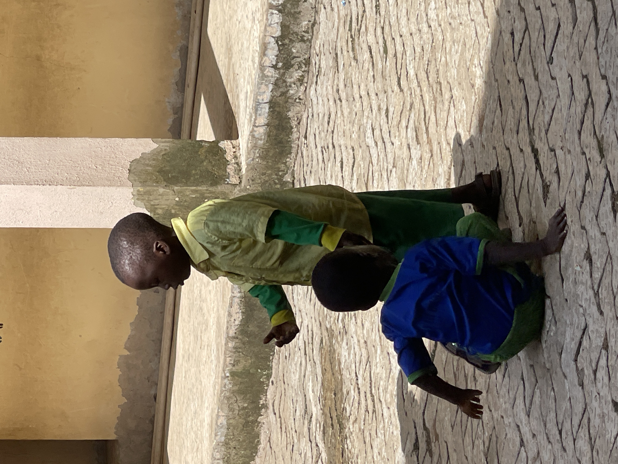 Two young boys are in an outdoor courtyard paved with interlocking bricks. The boy in the foreground is wearing a blue shirt with green trim and green pants, and he is crouched on the ground, facing away from the camera. His left arm is extended forward. The boy in the background is standing and wearing a light green short-sleeved shirt over a green long-sleeved shirt, and green pants. He is facing left and gesturing with his right hand. Behind the boys, a textured, pale yellow wall with a weathered, greenish-grey base runs along the back of the courtyard, with a white pillar to the left of it. The ground is partially shaded, indicating direct sunlight. The image appears to have been taken in Akwanga, Nigeria.