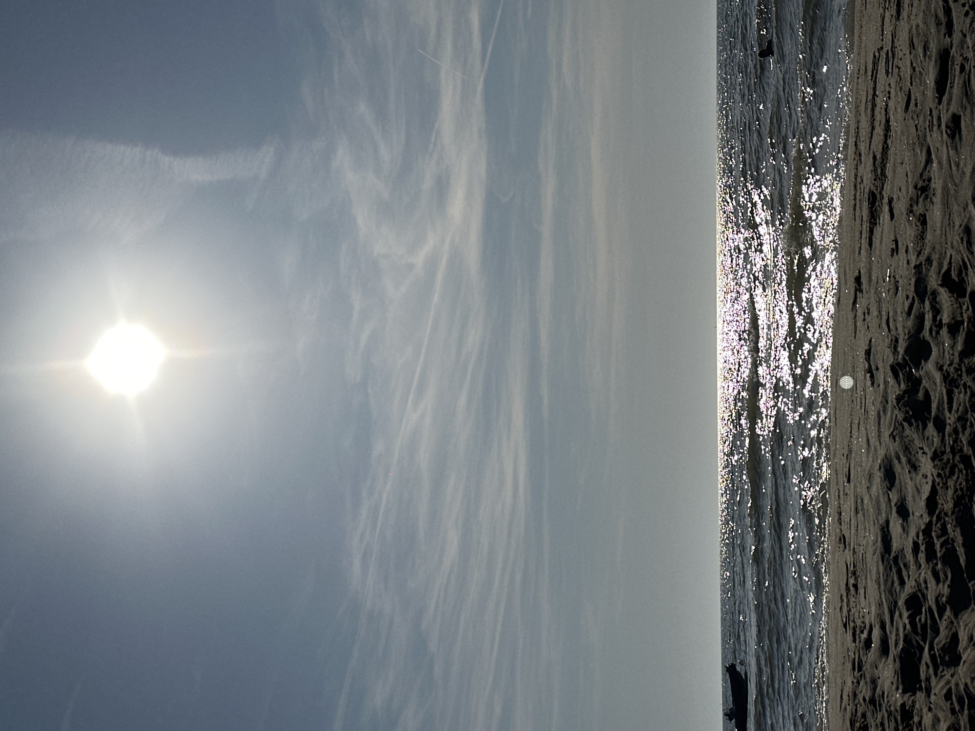 The image depicts a sunny day at the beach in Zandvoort, Netherlands. In the foreground, there is a sandy beach with textured patterns. The middle ground shows the ocean with waves and the sun reflecting brightly on the water's surface, creating a shimmering effect. To the left, a dark-colored boat is visible in the water, and to the right, a person is swimming. The background is dominated by a bright sun in the upper portion of the frame, partially obscured by hazy clouds. The sky is a pale blue with wispy clouds streaking across it, and faint contrails are visible in the upper right quadrant, suggesting the presence of aircraft. The overall scene conveys a bright and dynamic atmosphere of a typical beach day.