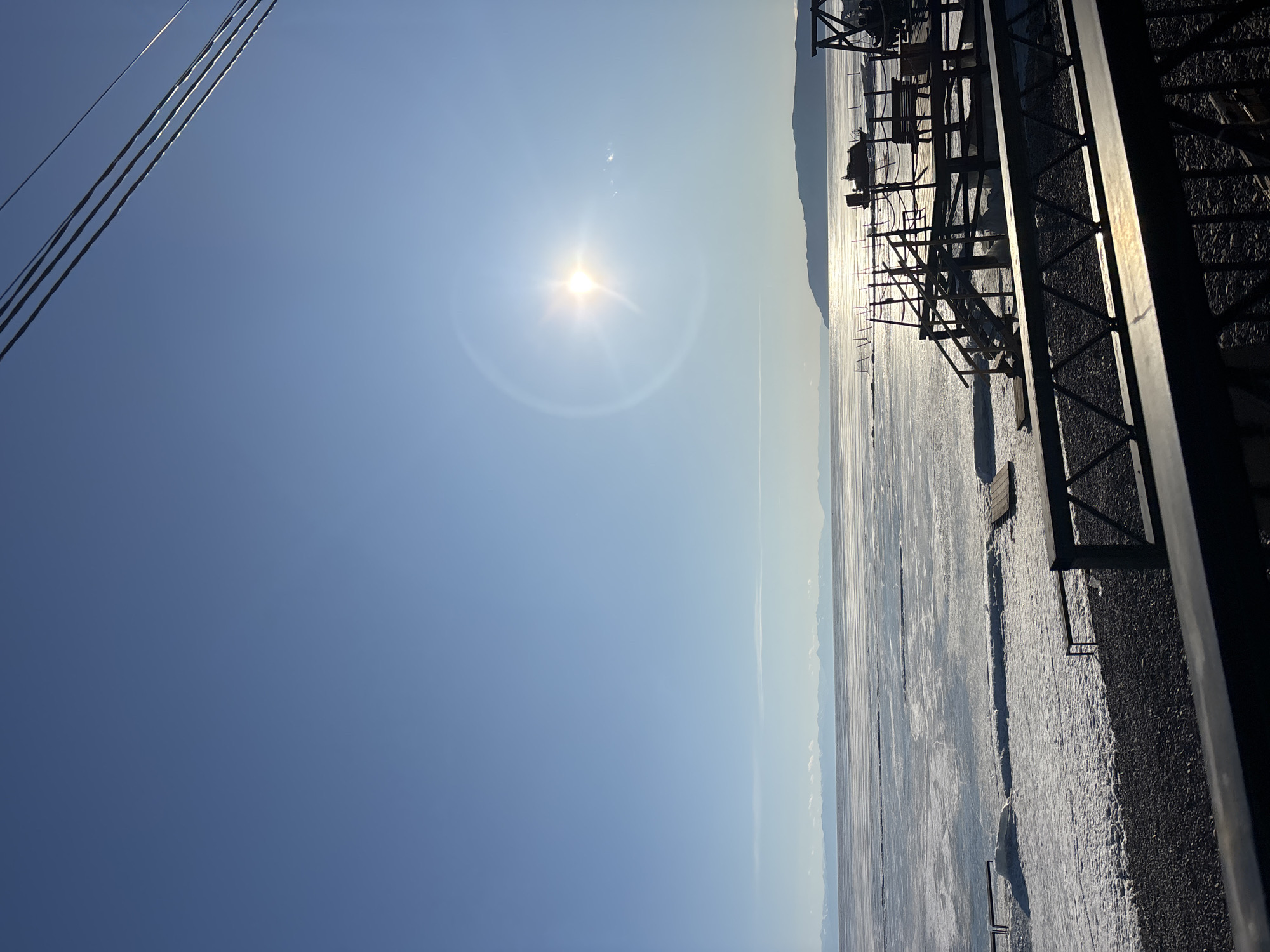 The image captures a panoramic view of a vast, partially frozen body of water, identified as Lake Baikal, from an elevated perspective in Listvyanka, Russia. The upper two-thirds of the frame consists of a clear, bright blue sky with a prominent, luminous sun positioned in the upper right quadrant, exhibiting a distinct lens flare. Multiple dark utility lines diagonally traverse the extreme upper right corner.
The horizon line separates the sky from a distant landscape featuring low-lying, snow-capped mountains to the left and a larger, dark landmass (likely a hill or mountain) to the right. The expansive middle ground is dominated by the frozen surface of Lake Baikal, which displays a varied texture of smooth ice sheets, fractured areas, and patches of accumulated snow.
The immediate foreground comprises a dark, pebbled or gravelly shoreline, transitioning into a mixture of ice and snow at the water's edge. Several wooden structures, consistent with docks or platforms, extend from the shore onto the frozen lake. On the right, a multi-tiered wooden platform features visible stairs and benches. A small, dark boat or amphibious vehicle rests on the ice near this structure. To the left, a simpler wooden dock or plank extends onto the ice. Several dark, slender posts are vertically embedded in the ice or shore in various locations.
At the bottom of the frame, a dark metal railing with a geometric lattice pattern occupies the immediate foreground, indicating the image was captured from a balcony or elevated viewing area. Indistinct figures appear to be seated on a bench located on the multi-tiered wooden platform to the right. The scene is brightly illuminated by direct sunlight, casting strong reflections across the ice and creating visible shadows on the foreground railing and wooden structures.