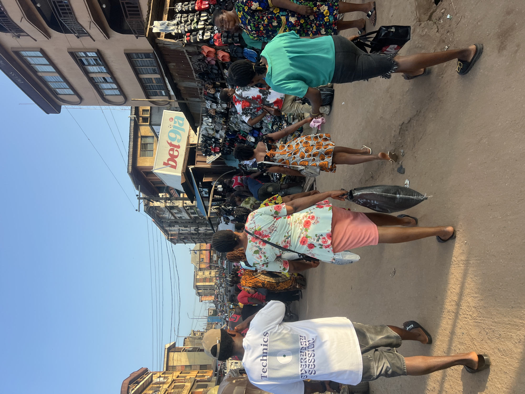 This is a medium shot photograph taken from behind a group of people walking away from the camera down a dusty, outdoor market street in Nkpor, Nigeria. The time of day appears to be late afternoon or early evening due to the low angle of the sun casting long shadows.
In the foreground, on the left, a man wearing a white t-shirt with "Technics" and "EVERYDAY SESSION" printed on the back, grey denim shorts, and black flip-flops walks away from the camera. He is wearing a light brown baseball cap. Next to him and slightly ahead is a woman wearing a light blue floral print dress with a pink skirt, carrying a large black plastic bag.
To their right and further down the street, another woman is walking away from the camera. She is wearing a short orange and white patterned dress and white flip-flops, carrying a light pink cloth. A woman in a bright teal t-shirt and dark shorts, wearing black slides, walks in the same direction. Further to the right, a woman in a colorful floral print dress with a yellow sash and dark sandals is visible.
The street is lined with buildings on both sides. The buildings are multi-story structures with a mixture of concrete and corrugated iron. On the right side of the street, stalls are set up displaying numerous pairs of shoes and sandals. A prominent sign for "bet9ja" is visible above one of the stalls. The street itself is unpaved and appears to be a dirt or sand surface. In the background, the street continues, filled with more people and what appears to be traffic congestion. The sky is clear and blue.