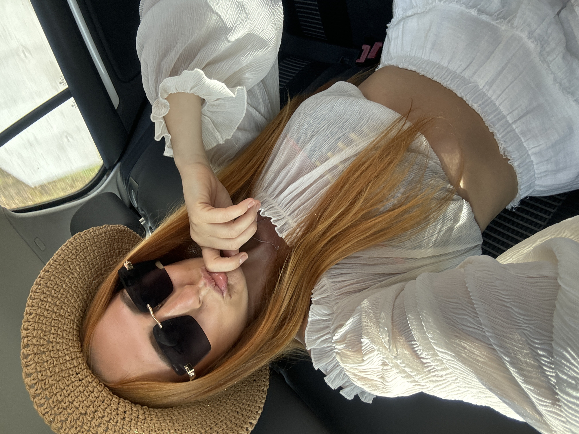 A female individual with long, reddish-orange hair is depicted in a selfie, seated inside the passenger compartment of a vehicle, positioned in Budva, Montenegro. She wears a wide-brimmed, light brown woven straw hat and dark-lensed, rectangular sunglasses with metallic accents. Her attire consists of a sheer, white, crinkle-textured crop top with voluminous, ruffled sleeves, exposing her midriff, paired with a matching white, ruffled, tiered skirt or shorts featuring an elasticated waistband. Her right hand, with natural-looking fingernails, is raised, with the index finger touching her puckered lower lip, indicating a deliberate pose. The vehicle's interior features black and grey upholstery and trim elements, including a visible headrest and a door panel with a silver accent strip. Bright sunlight enters through a rear window, creating strong highlights and reflections. Blurred green and yellow foliage is visible through the window, suggesting an outdoor, possibly natural, landscape beyond the vehicle.