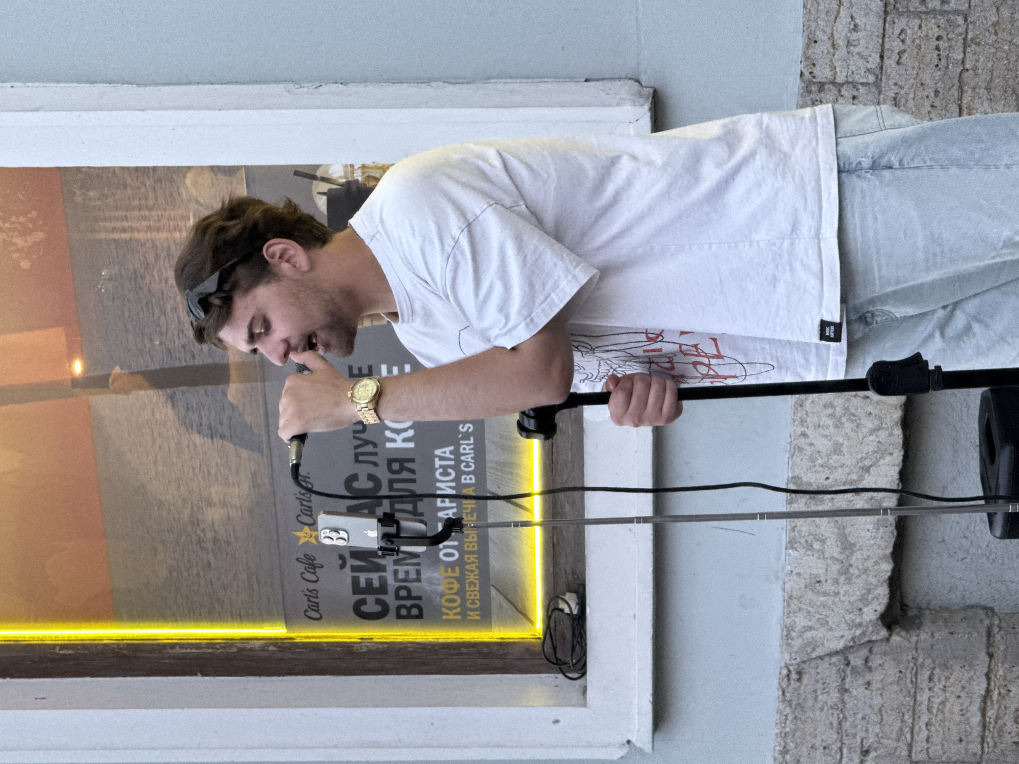 A young man with brown hair, wearing sunglasses perched on his head, a white t-shirt, and light blue jeans, leans forward, singing into a microphone. He is holding the microphone with his right hand and resting his left arm on a black stand. A gold watch adorns his left wrist. He appears to be performing outdoors, as indicated by the natural light and the textured stone wall in the background.
Behind the man, a window displays a poster with Cyrillic text and an image of a body of water. A bright yellow neon light runs along the bottom of the window frame. Visible text on the poster includes "Carl's Cafe Carls," "СЕЙ АС ЛУЧЕ," "ВРЕМ ДЛЯ КО," "КОФЕ ОТ АРИСТА," and "И СВЕЖАЯ ВЫЛЕЧКА В CARL'S." A smartphone is mounted on a stand in front of the microphone, suggesting he might be recording or live-streaming his performance. The scene is likely taking place during the day, given the bright lighting. The man's focused expression suggests he is engrossed in his singing. The overall setting appears to be an urban environment, possibly a cafe or a street with a commercial establishment.