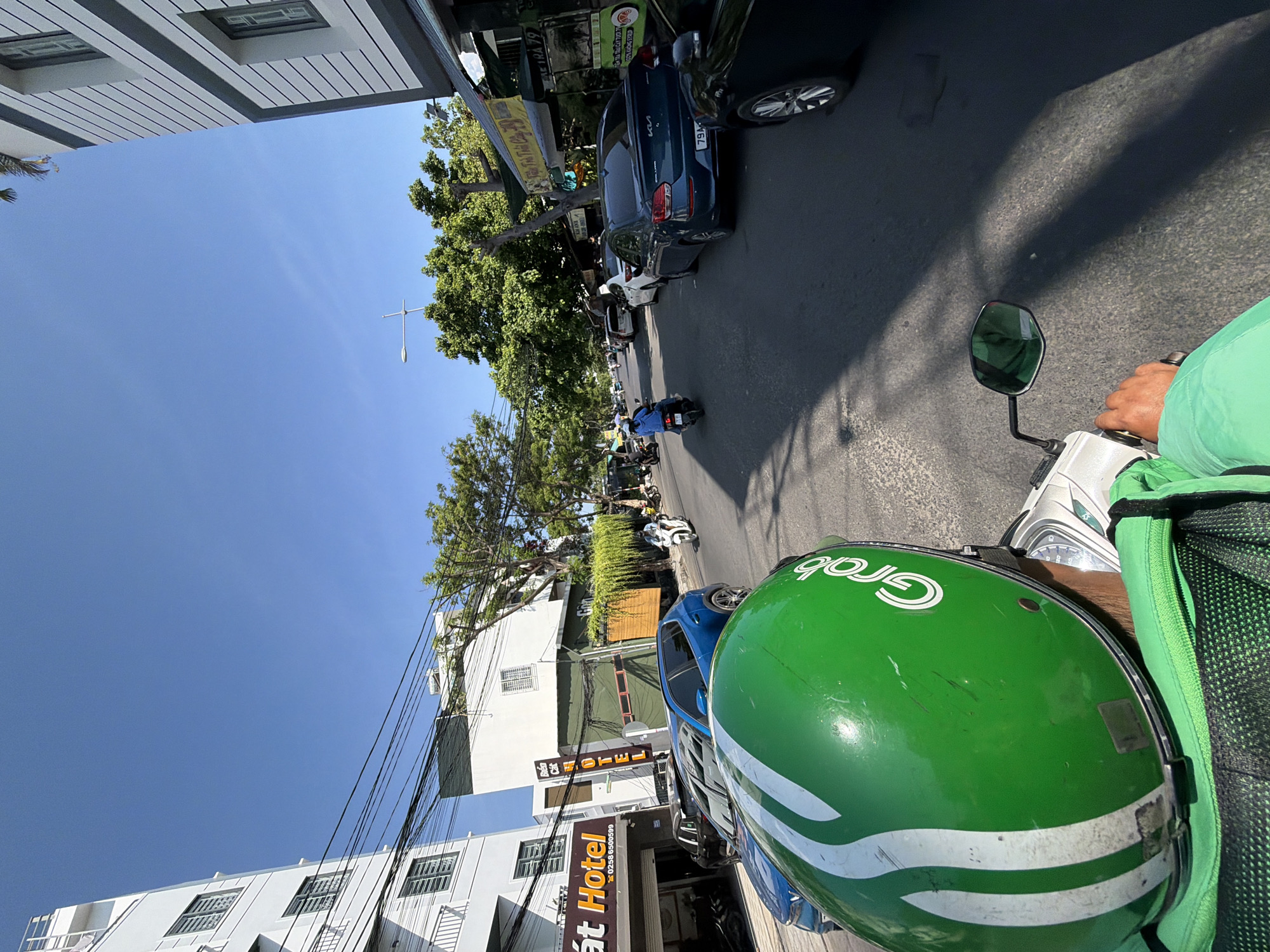 A POV shot from a motorcycle helmet in the city of Nam Nha Trang Ward, Vietnam. The helmet is green with white stripes and the "Grab" logo. The rider is wearing a green jacket. The motorcycle's right side mirror is visible. The road is asphalt and lined with buildings and trees. There are several cars and motorcycles on the road. The sky is clear blue. A hotel sign with "Dat Hotel" is visible on the left.