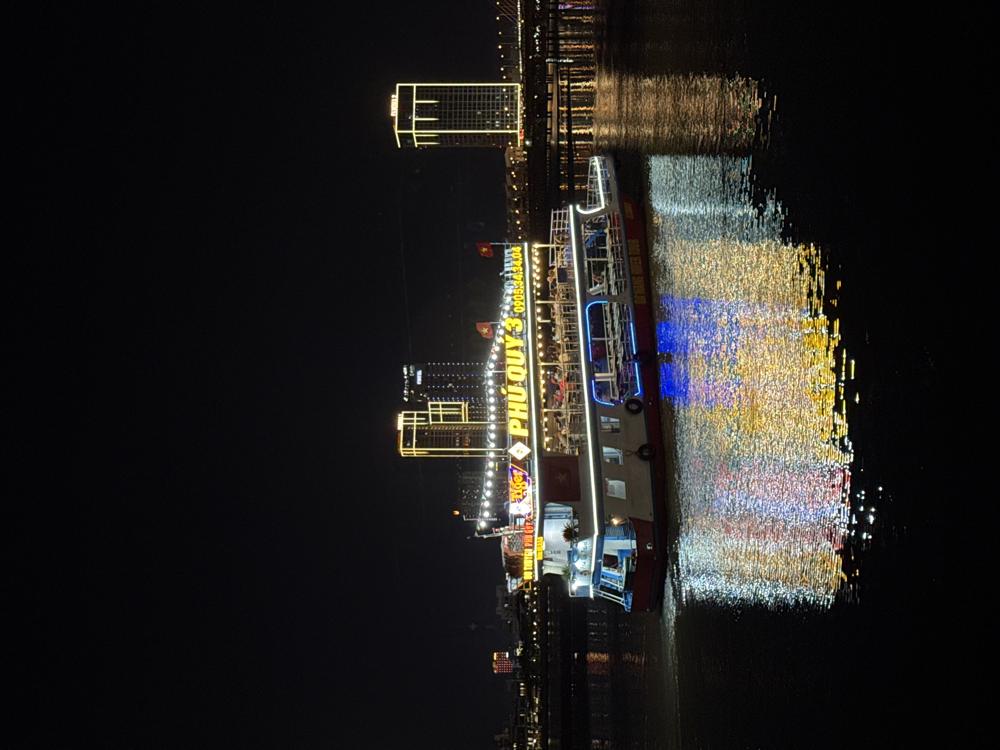 The image captures a vibrant night scene in Đà Nẵng, Vietnam, centered around a brightly illuminated passenger boat cruising on a calm river. The boat, named "PHU QUY 3" in large yellow letters, dominates the midground, adorned with numerous lights, including prominent blue and yellow strips, creating a festive atmosphere. Below the name, a phone number "0905.34.34.04" is visible. Two Vietnamese flags are also seen flying from the boat.
People are visible on the boat's multiple decks, appearing to be passengers enjoying a night excursion. In the background, a modern city skyline is visible, with several buildings lit up. A tall building with distinct yellow outline lights stands out prominently. The artificial lights from both the boat and the city reflect dazzlingly on the water's surface, creating a shimmering, colorful effect that includes hues of blue, yellow, and white. The time of day is clearly night, with the dark sky providing a stark contrast to the brilliant lights. The scene suggests a popular tourist activity or an evening of leisure on the river, possibly a dinner cruise.