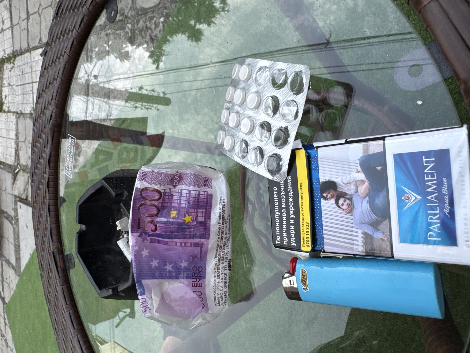 The image captures a casual arrangement of items on a glass-topped outdoor table, likely on a patio in Varna, Bulgaria. The table has a dark, woven rattan-style edge and reflects greenery, suggesting a daytime, open-air setting.
At the center of the composition is a light blue BIC lighter next to a white and blue pack of Parliament Aqua Blue cigarettes. The cigarette pack features an image of a man and a woman and displays a health warning in Cyrillic: "Тютюнопушенето причинява мозъчни удари и увреждания" (Smoking causes strokes and injuries), along with a helpline: "Помощ за отказ от тютюнопушене: 0700 10 323".
To the left, a black ashtray is visible, adjacent to a clear plastic bag containing a crumpled 500 Euro banknote. The plastic bag bears text indicating its origin from "JUMBO S.A." in Athens, Greece, with "Made in China" and various translations for "produced in China" and "imported by." Above these items lies an empty silver blister pack, partially still holding several white, round pills, while many slots are empty.
The collection of objects—cigarettes, lighter, ashtray, pills, and cash—suggests a moment of leisure or a casual outdoor gathering. The "TEMPERED GLASS KRAFT-WORLD.BG" sticker on the table's surface identifies the furniture. The presence of the Euro banknote and Cyrillic text on the cigarette pack aligns with the provided location context of Bulgaria.