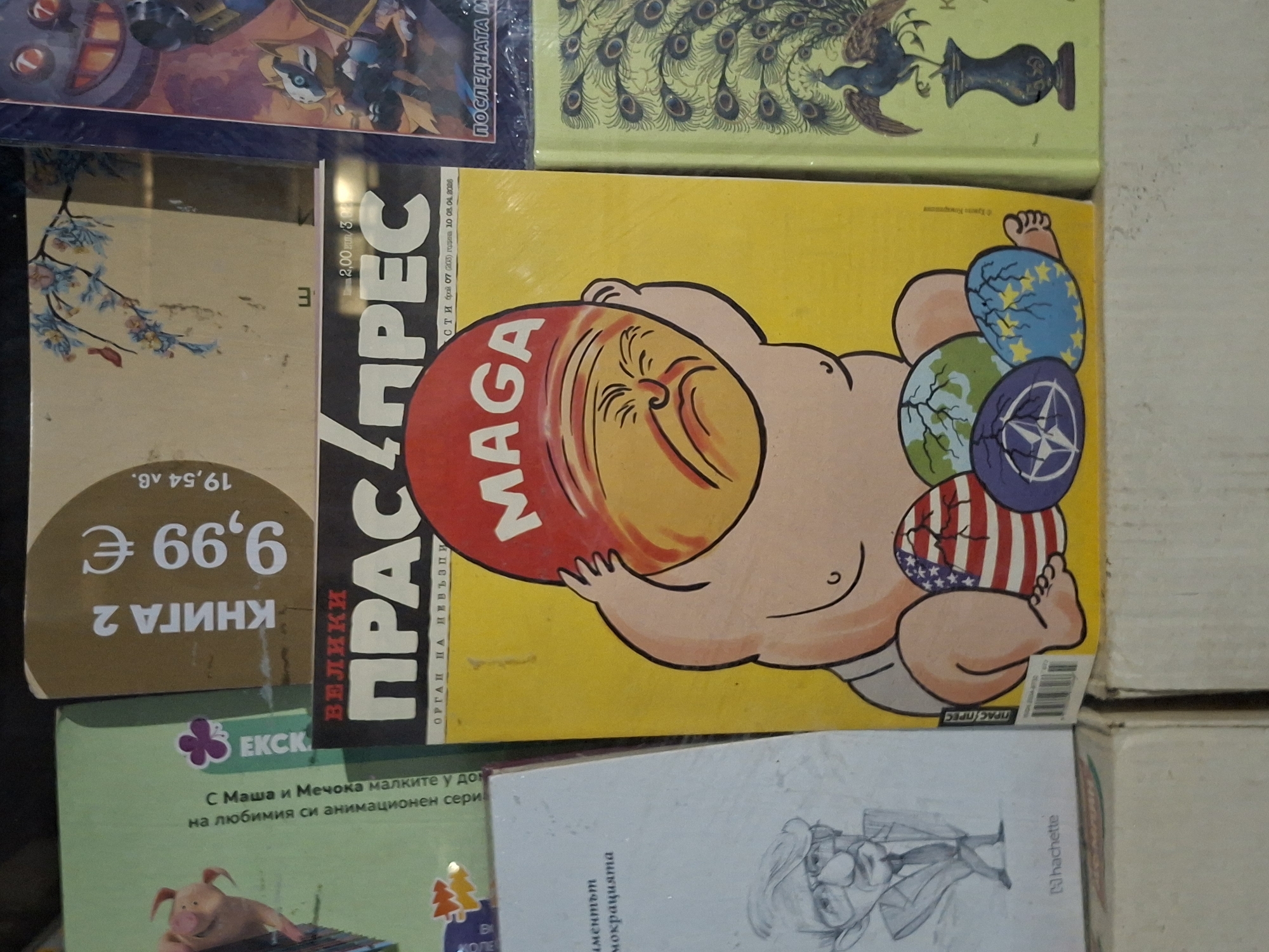 The image shows a stack of books and magazines, including a satirical magazine cover with a cartoon of a baby wearing a MAGA hat and holding spheres decorated with the EU flag, the NATO symbol, and the US flag. The magazine is titled "ПРАС ПРЕС" ("PRAS PRES"), which loosely translates to "Pig Press." The price of another book in the stack is displayed as "9,99 €" and "19,54 лв.", indicating a location in Bulgaria, where both currencies are commonly used. The Bulgarian text on the magazine cover confirms the location. Given the prominence of Bulgarian text and the fact that both euros and Bulgarian lev are used, it can be assumed the image was taken in Plovdiv, Bulgaria.