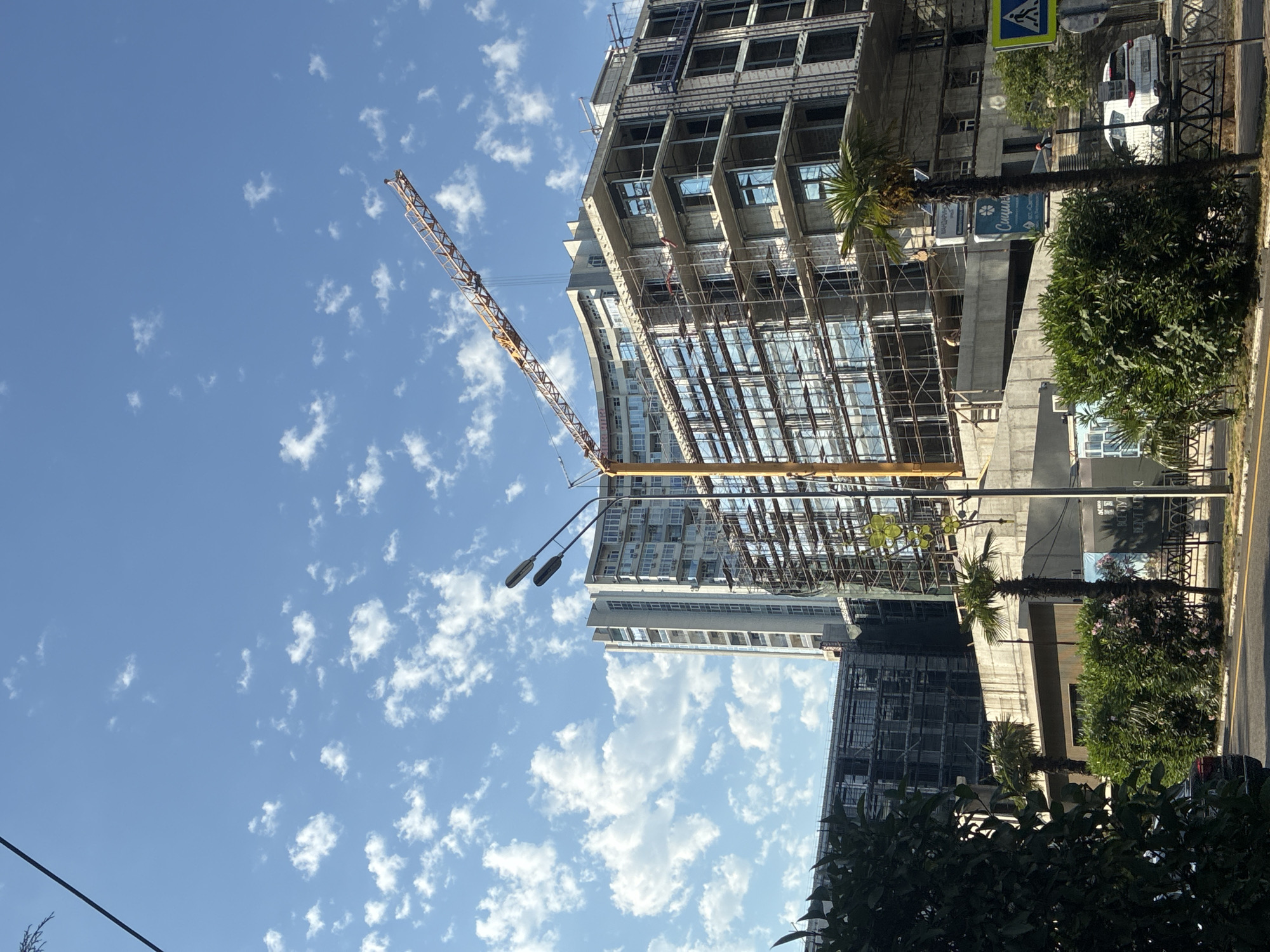 The image shows a construction site in Sochi, Russia, on a sunny day with scattered clouds. A large, multi-story building is under construction, covered in scaffolding. A yellow construction crane is prominently visible, extending over the building. In the foreground, there are trees and bushes, and a street with a white car and a pedestrian crossing sign. One of the buildings under construction has windows that reflect the blue sky and clouds. There are glimpses of other buildings in the background. A sign with Cyrillic text is visible on one of the structures.