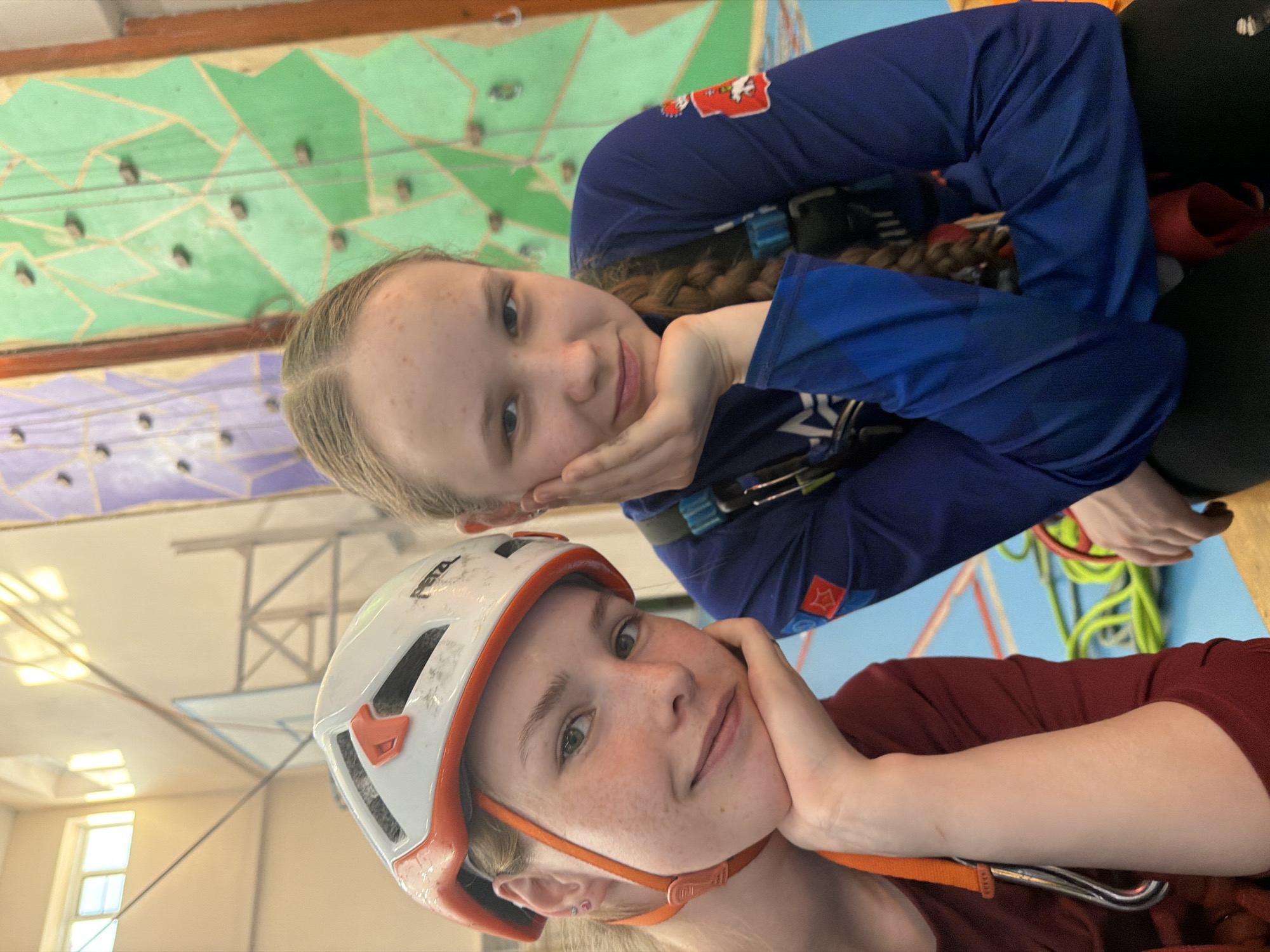 Two young women are posing indoors. The woman in the foreground is wearing a white Petzl helmet with orange trim and a reddish-brown shirt. Her left hand is supporting her chin. The woman in the background is wearing a blue long-sleeved shirt with a red and white patch on the left sleeve, a climbing harness, and has her hair in a braid. Her left hand is also supporting her chin. Behind them is an indoor rock climbing wall with green and purple sections. Climbing equipment such as ropes are visible in the background. A window is visible in the background, and a metal support structure. The location is Berezniki, Russia, confirmed by the patch on the woman's sleeve.