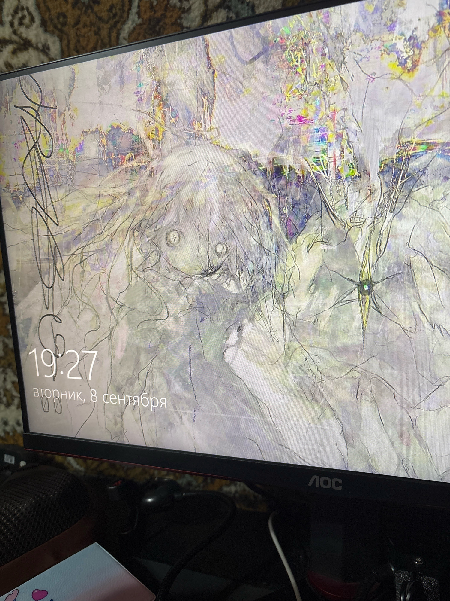 The image shows a computer monitor displaying a lock screen with a time of 19:27 and the date "вторник, 8 сентября" (Tuesday, September 8). The wallpaper is an abstract, sketch-like illustration with hints of color. The monitor has an "AOC" logo at the bottom. The background behind the monitor appears to be a patterned carpet. The scene is likely indoors, possibly in a home or office. There is no visible indication of the city of Novocheboksarsk, Russia, or any specific location within it.
