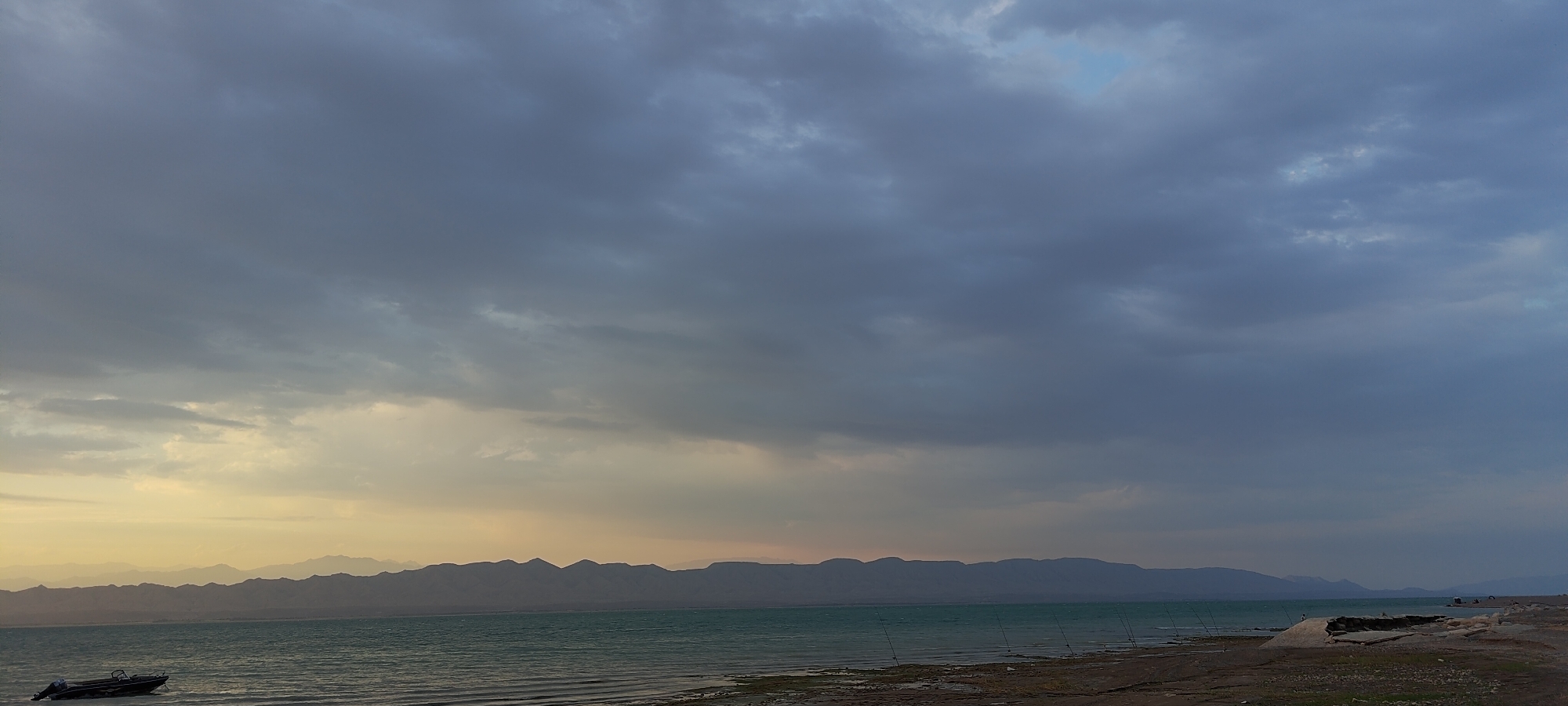 The image shows a body of water with a distant mountainous shoreline under a cloudy sky. The water is a turquoise green, and the shore is rocky and appears to be composed of sand and pebbles. Several thin, vertical poles, likely fishing rods, are scattered along the water's edge. In the foreground, a small, dark boat is partially submerged near the left edge of the frame. The sky is filled with heavy, dark gray clouds, with patches of lighter, yellowish light breaking through near the horizon. The mountains in the distance are silhouetted against the sky, with layered peaks stretching across the frame. The overall scene suggests a natural landscape, possibly at dusk or during overcast weather. No people are clearly visible in the image.
