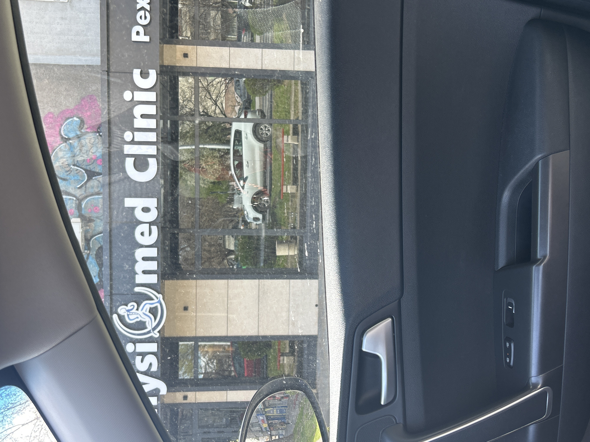 The image, taken from inside a car, shows a view of a building across the street, reflected in the window. The building has a sign that reads "Physimed Clinic Pex" vertically. There is graffiti on the side of the building. A white car is visible in the reflection of the windows. The image appears to be taken in an urban environment, potentially Plovdiv, Bulgaria, given the clinic's name. The car's interior, including the door panel with its handle and window/lock controls, is visible in the foreground. The scene is taken during daylight. No people are discernible within the reflection. The overall environment suggests a commercial or urban area.