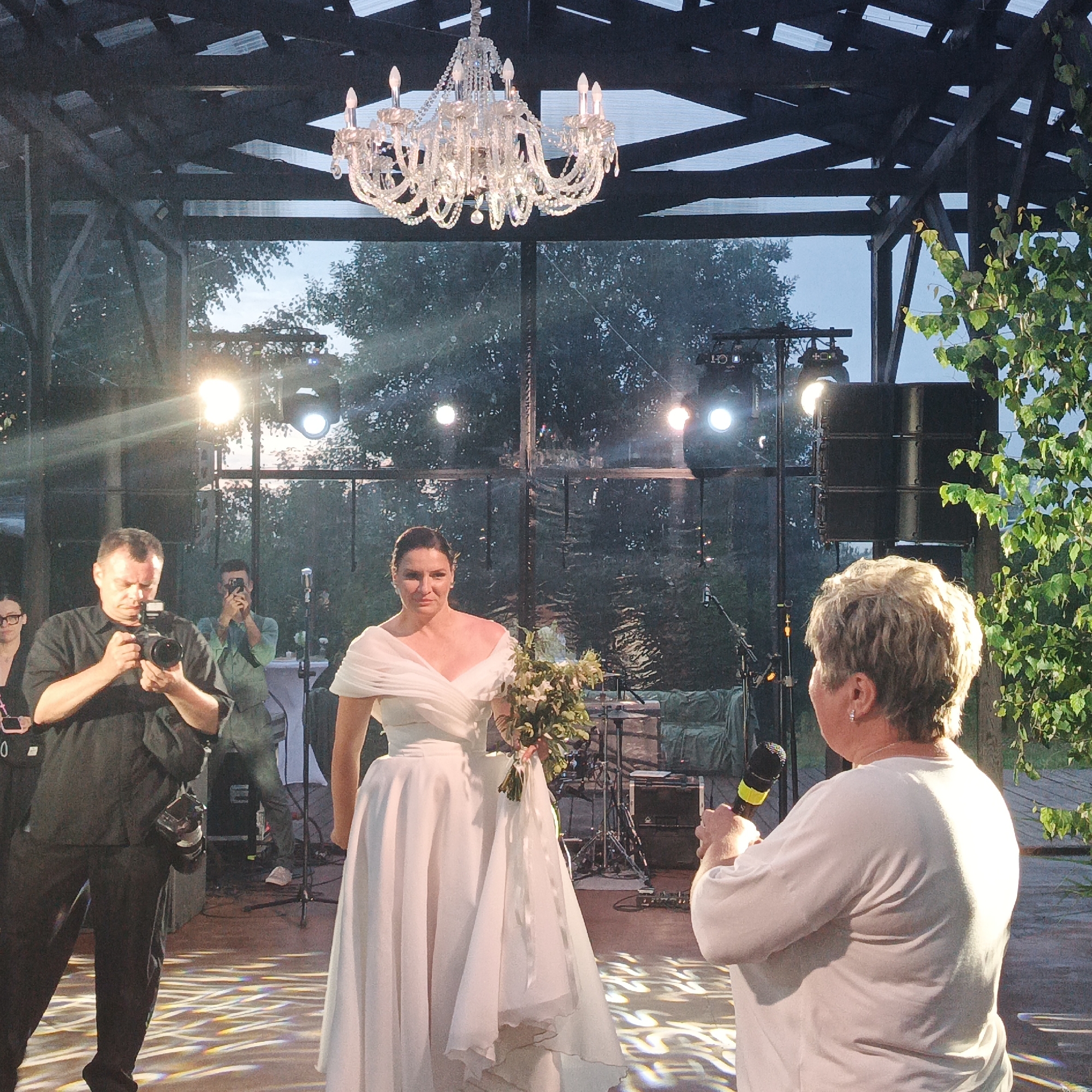 The image captures an outdoor event, possibly a wedding reception, at the "Svetloye" restaurant complex in Svetloye, Russia. In the foreground, a woman in a white wedding dress stands holding a bouquet of greenery. To her left, a man in a black shirt and pants is photographing her with a professional camera. Behind the bride, another man is seen taking a photo with his phone. In the mid-ground, a speaker system and stage lighting are visible. The background features trees and the sky, indicating an evening or late afternoon setting. The overall atmosphere appears festive, with a large crystal chandelier hanging from the structure above.