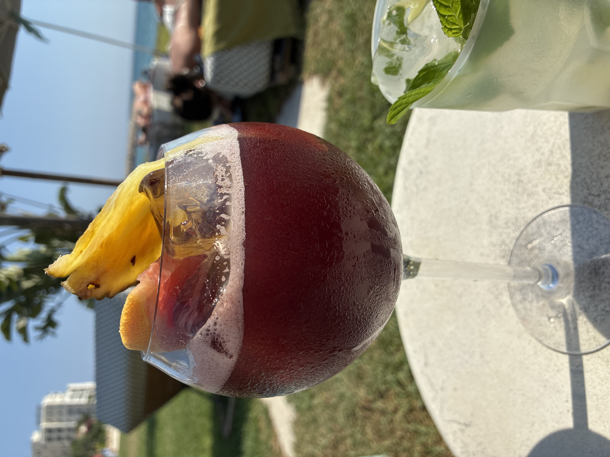 In Limassol, Cyprus, on a sunny day, a close-up shot focuses on a tall glass filled with a dark red beverage, garnished with a slice of pineapple and a segment of citrus fruit. The glass is on a white table, with condensation visible on its surface, indicating it is a cold drink. To the right, a frosted glass containing a lighter colored drink with ice and mint leaves is partially visible. In the background, a grassy area leads towards a blurred scene of people relaxing on lounge chairs under umbrellas, with a view of the Mediterranean Sea and distant buildings. The overall ambiance suggests a relaxing day at a beachside resort.