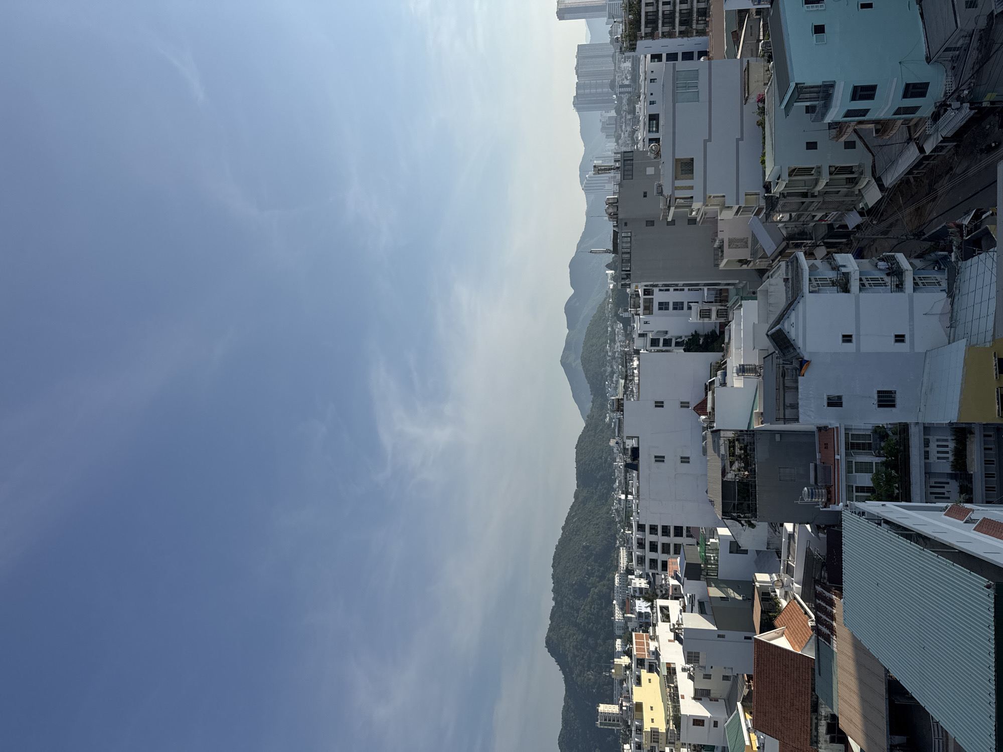 The image captures a daytime aerial view of a densely populated urban area in Nha Trang, Vietnam, with a backdrop of lush green mountains and a clear blue sky. The foreground is dominated by a mix of residential and commercial buildings of varying heights and architectural styles, featuring white, yellow, and blue facades, some with terracotta or green metal roofing. The buildings are tightly packed, with narrow streets and alleyways visible between them, crisscrossed with overhead power lines. In the mid-ground, a cluster of taller, modern buildings, possibly apartment complexes or hotels, stand out. The background is characterized by rolling, tree-covered hills that extend to the horizon, suggesting a coastal city setting. The lighting indicates it is likely morning or late afternoon, with soft sunlight casting subtle shadows. No individuals are clearly identifiable in the image.