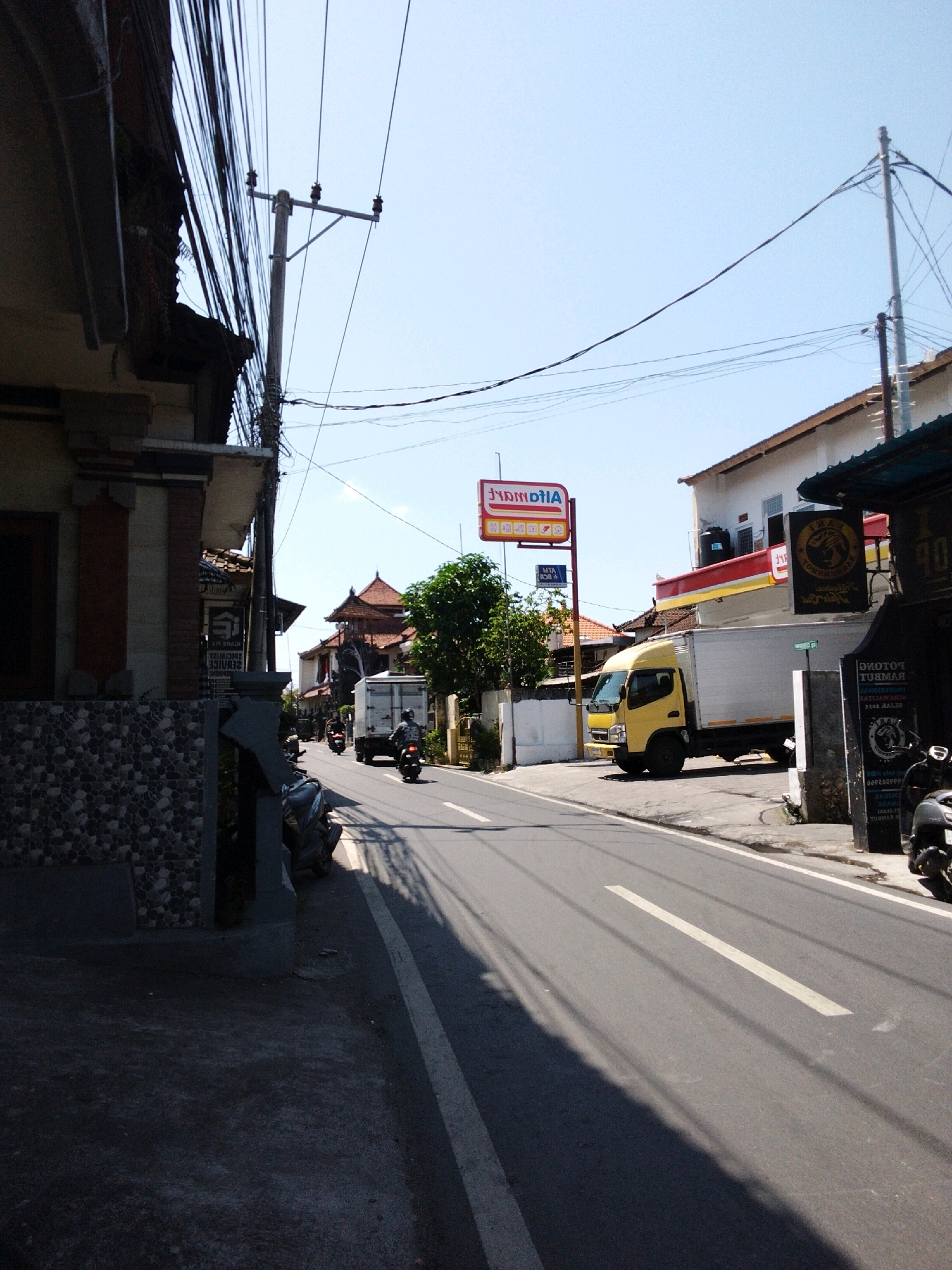 The image depicts a street scene in Denpasar, Indonesia. On the left, a building with a tiled facade and dark wooden eaves is partially visible. Overhead, a dense network of electrical wires hangs from a utility pole. The street itself is paved with asphalt and marked with white lines. A yellow box truck with a white trailer is parked on the right side of the road. In front of the truck, a red and yellow sign for "Indomaret" is visible, indicating a convenience store. Several motorcycles are parked along the side of the road. Further down the street, traditional Balinese architecture is evident in the buildings with tiled roofs. The sky is clear and blue.