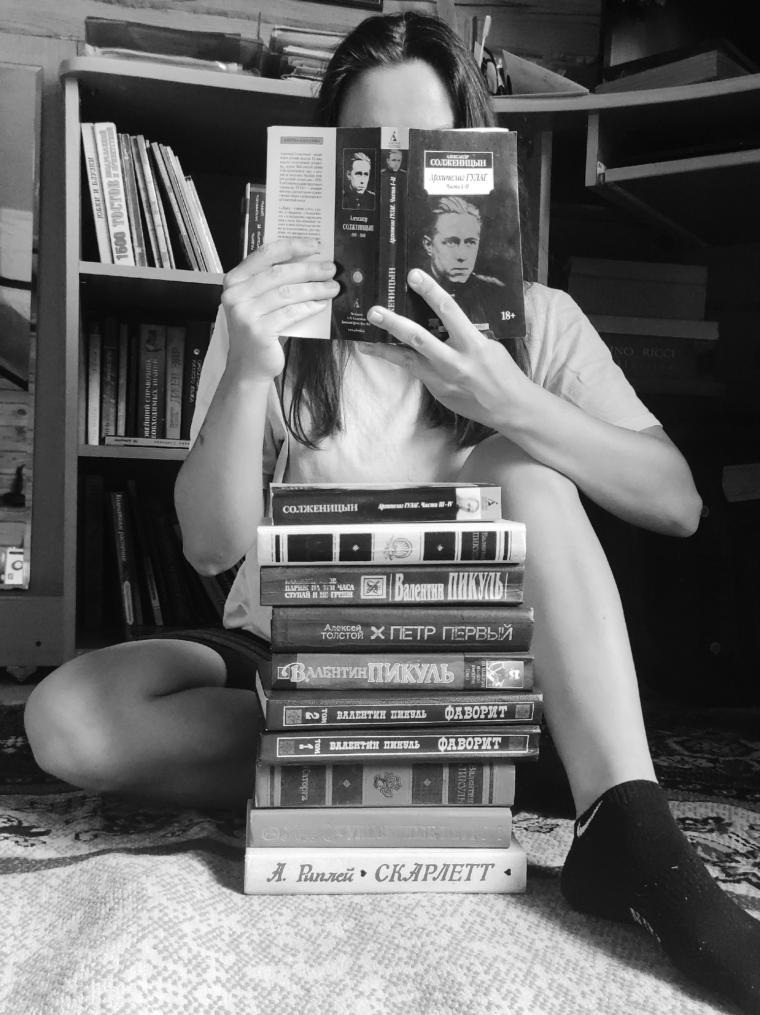 A black and white photograph captures a person sitting on a patterned rug, engrossed in reading. The individual's face is obscured by the book they are holding open, revealing only parts of their arms, legs, and torso. They are wearing a light-colored t-shirt, dark shorts, and one black Nike sock is visible on their right foot.
In front of the person is a tall stack of books, and behind them, a bookshelf filled with more books lines the wall. The book being read is titled "Архипелаг ГУЛАГ. Части I-II" (The Gulag Archipelago. Parts I-II) by Александр СолженицыН (Aleksandr Solzhenitsyn), prominently displaying an "18+" age rating and a portrait of the author on its cover.
The stack of books includes:
- "СОЛЖЕНИЦЫН Архипелаг ГУЛАГ. Части III-IV" (Solzhenitsyn The Gulag Archipelago. Parts III-IV)
- A book by Валентин ПИКУЛЬ (Valentin Pikul), possibly "Реквием караванам PQ-17" (Requiem for Convoys PQ-17)
- "ВАЛЕНТИН ПИКУЛЬ ПАРИЖ НА ТРИ ЧАСА СТУПАЙ И НЕ ГРЕШИ" (Valentin Pikul Paris for Three Hours, Go and Don't Sin)
- "Алексей ТОЛСТОЙ • ПЕТР ПЕРВЫЙ" (Alexei Tolstoy • Peter the First)
- "ВАЛЕНТИН ПИКУЛЬ" (Valentin Pikul), with the title "На задворках великой империи" (In the Backyards of the Great Empire)
- Two volumes of "ВАЛЕНТИН ПИКУЛЬ ФАВОРИТ" (Valentin Pikul Favorite)
- "А. Риплей • СКАРЛЕТТ" (A. Ripley • Scarlett)
Visible on the bookshelf are spines of various books, including "ЮБКИ И БЛУЗКИ" (Skirts and Blouses), "1500 ТОСТОВ" (1500 Toasts), "КОМНАТНЫЕ РАСТЕНИЯ" (Houseplants), and "ВАЖНЕЙШИЙ СПРАВОЧНИК НЕОБХОДИМЫХ ЗНАНИЙ" (The Most Important Handbook of Essential Knowledge). The scene conveys a quiet, personal moment of reading in what appears to be a home library or study space.