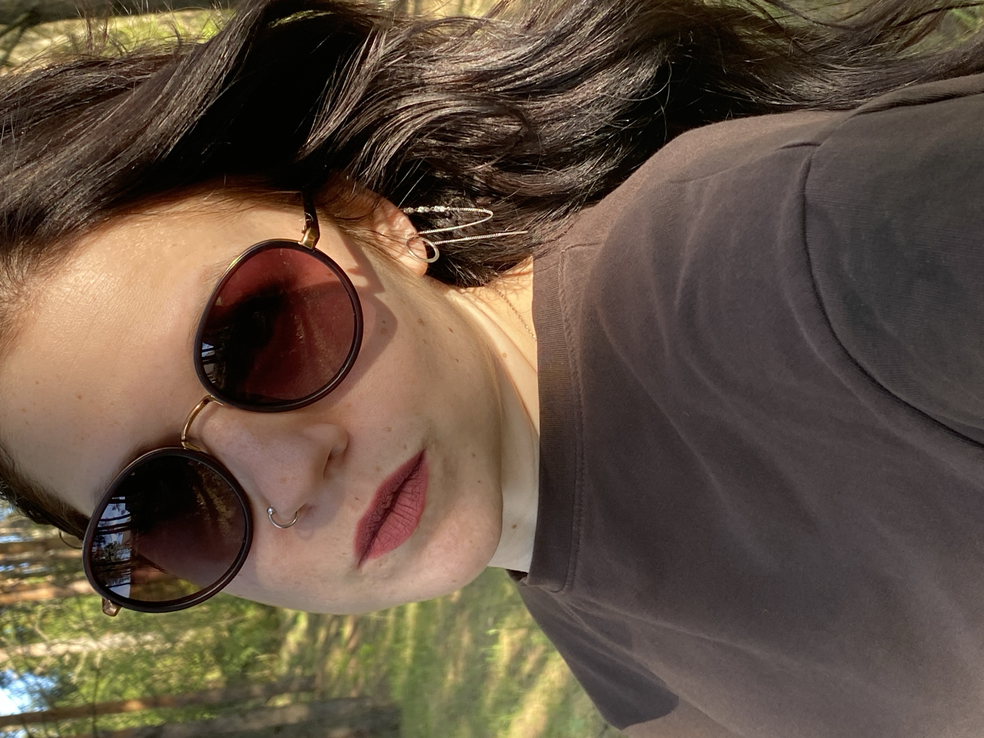 A woman with dark hair is wearing sunglasses and a dark-colored t-shirt. She has a nose ring and decorative ear jewelry. The background appears to be a forest or park setting, judging by the glimpses of green foliage and tree trunks visible through the reflection in her sunglasses. It is impossible to verify or deny that the image was taken in Фоки, Russia, from the image itself.