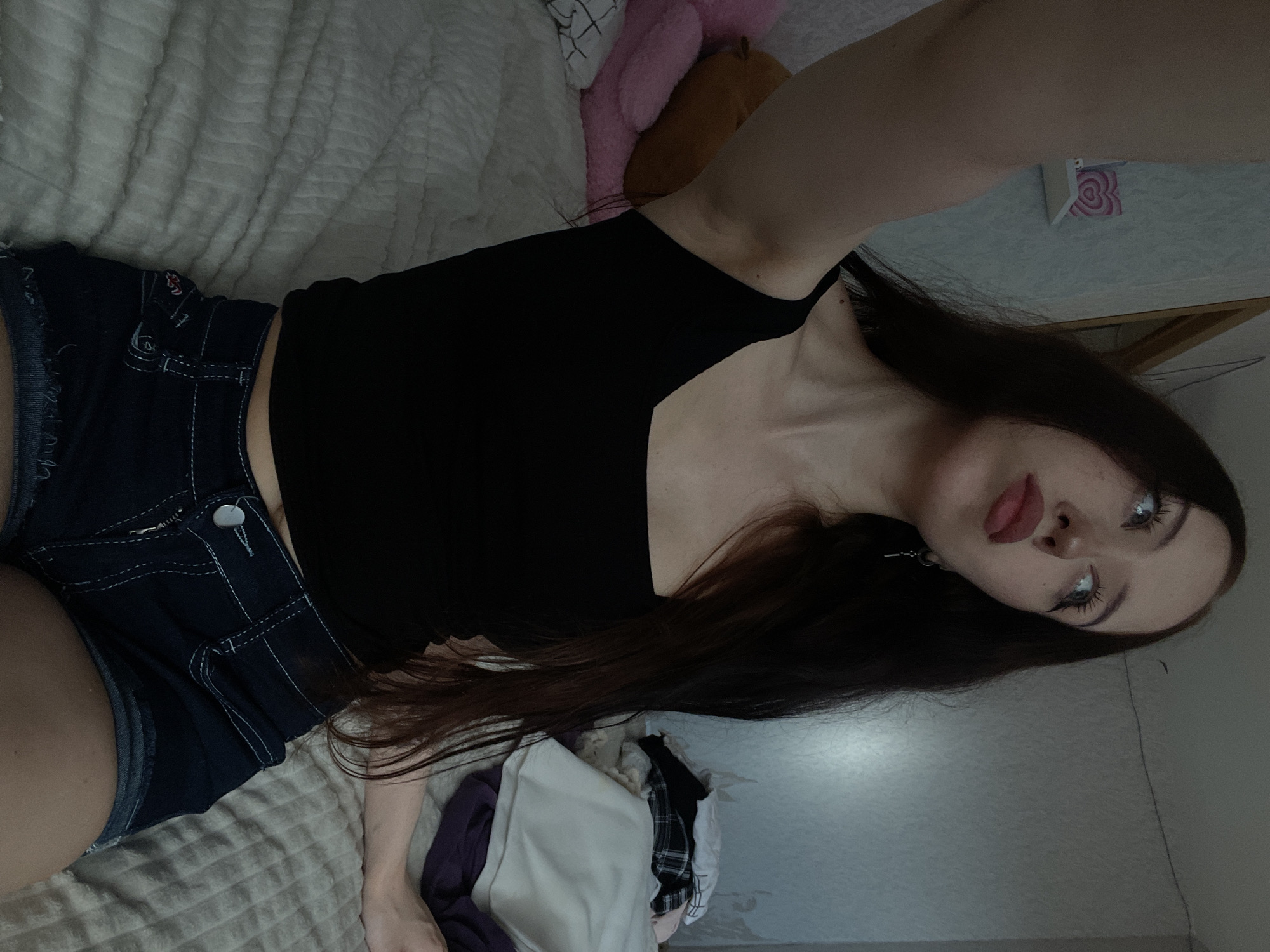 A young woman with long, dark brown hair is sitting on a bed, holding a phone to take a selfie. She is wearing a black tank top and denim shorts. The room appears to be a bedroom, with a bed, a shelf on the wall, and a pile of clothes in the background. The wall is a light blue color. The lighting in the room is dim, suggesting it is nighttime or the blinds are drawn. The overall atmosphere is casual and relaxed.