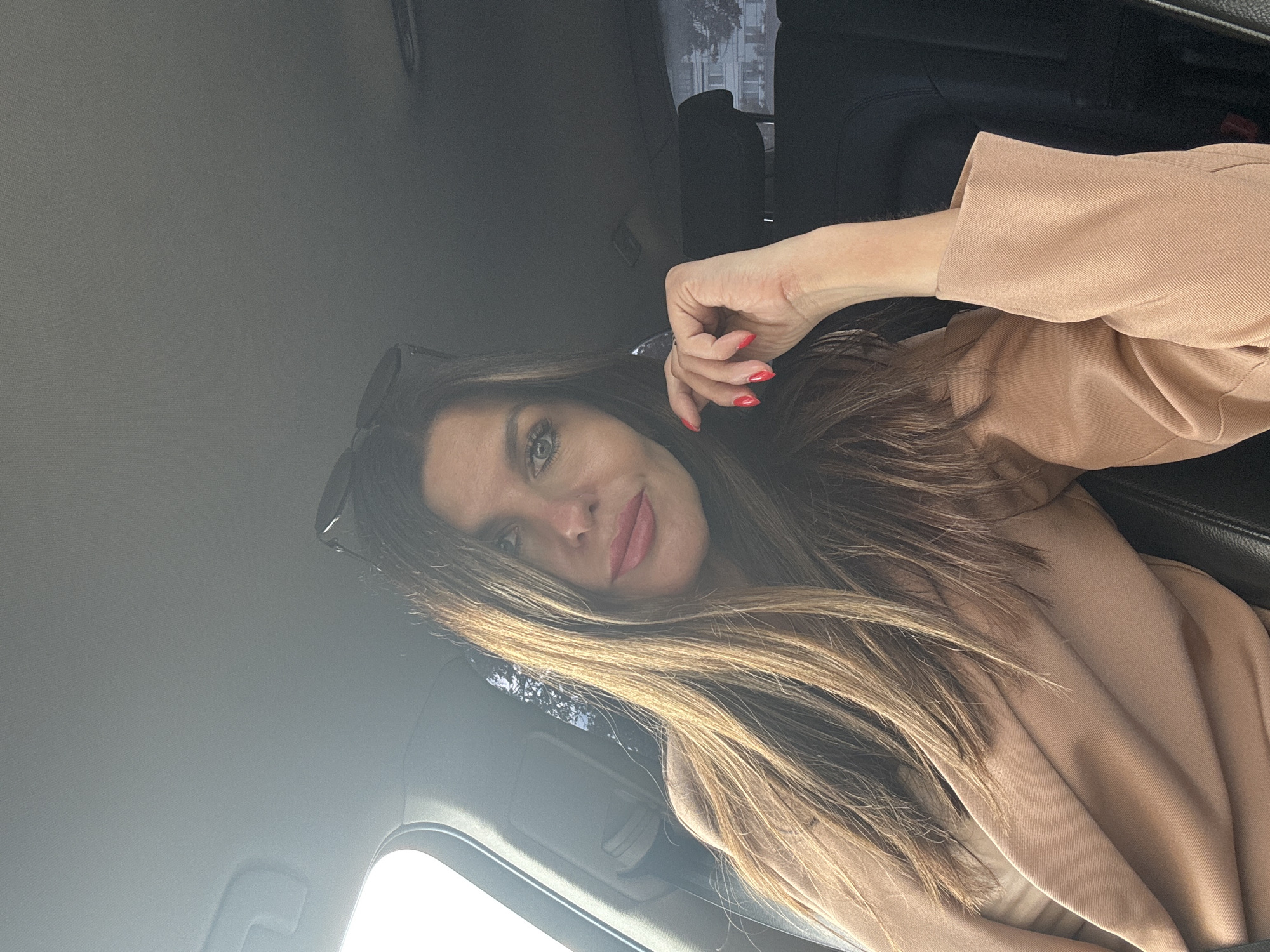 The image shows a woman sitting in the driver's seat of a car. She has long, brown hair and is wearing a beige jacket. Her right hand is resting on her chin, and her nails are painted red. Sunglasses are perched on top of her head. The interior of the car is visible, including the headrest of the passenger seat and the ceiling. Sunlight is streaming in from the left side of the car, casting shadows on the woman's face. The background outside the car shows a building with windows and trees, suggesting an urban or suburban environment. There is no specific visual information in the image to indicate the location is the city of Chaikovsky, Russia.