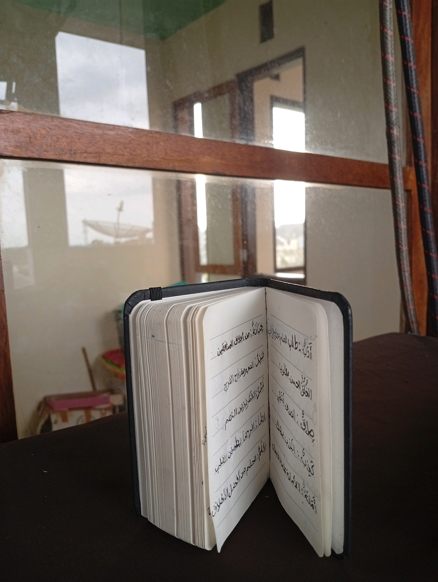 An open book with Arabic script is placed on a dark surface. The book has a black cover and thin white pages filled with handwritten text. The background reveals a window with reflections of a room and the outdoors. In the reflection, a satellite dish is visible, suggesting an outdoor setting. The overall scene appears to be indoors, with a window providing a view of the outside, possibly a residential area. The lighting suggests it is daytime.