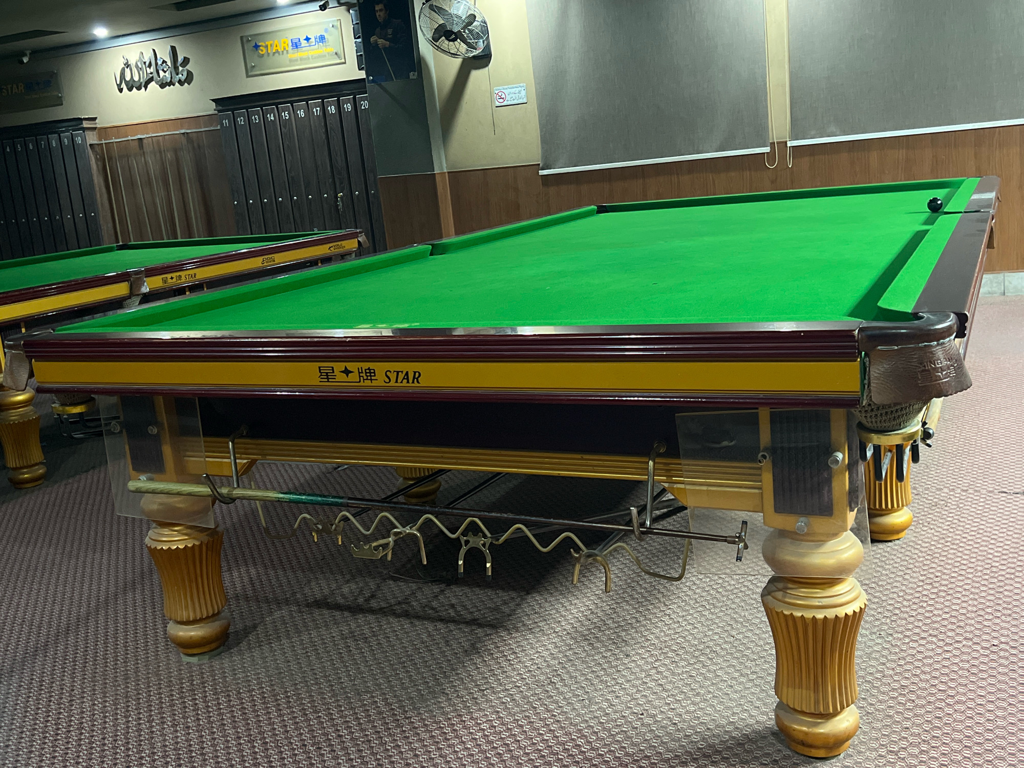 The image depicts a snooker hall in Attock, Pakistan, featuring multiple snooker tables. The foreground prominently displays a snooker table with a green felt surface, a wooden frame with maroon and yellow accents, and ornate carved wooden legs. A single black snooker ball rests on the table. Behind this table, another snooker table is partially visible. The background reveals a wall with various posters and signs, including one with Arabic script and another with Chinese characters and the word "STAR". A rack for snooker cues is attached to the side of the main table. The flooring is a patterned carpet. The lighting suggests an indoor setting, likely a commercial establishment.