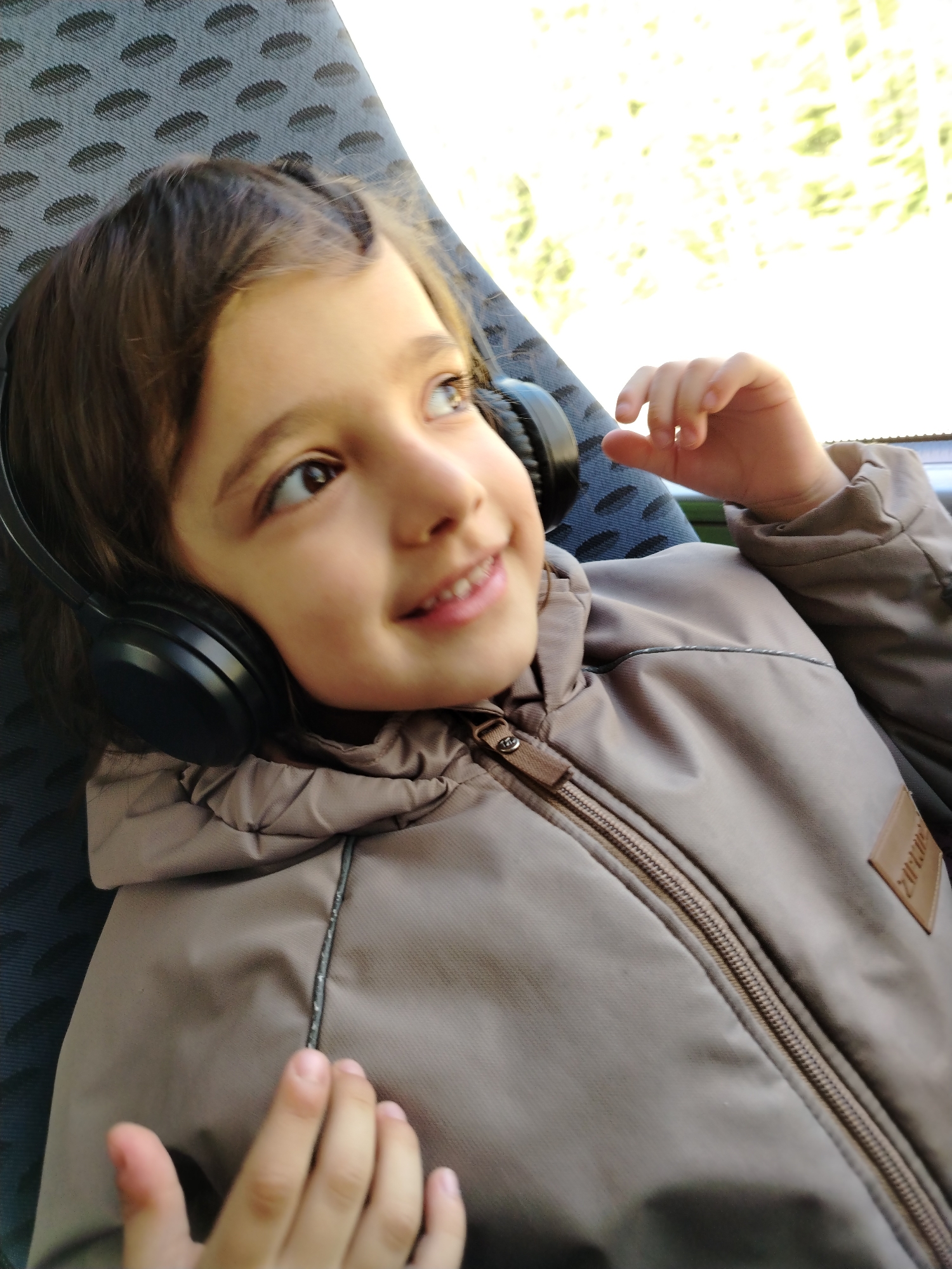 A child is reclining in a seat, possibly on a bus or train. They are wearing headphones and a zippered, hooded jacket. The child is looking out of a window, appearing to be smiling slightly. The seat has a patterned fabric. Based on the prompt, the location is Ol'khovochka, Russia.