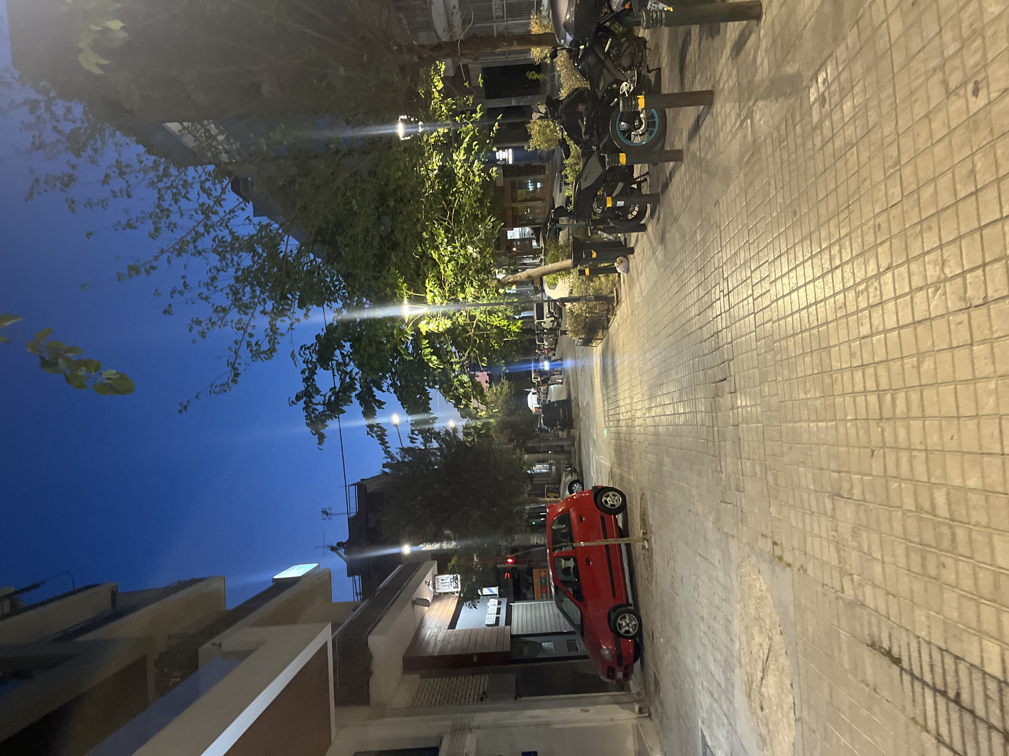 The image depicts a street scene in Athens, Greece, at dusk or early evening. The sky is a deep blue, transitioning to lighter shades near the horizon. The street is paved with square tiles, and sidewalks line both sides.
On the left side of the street, a red hatchback car is parked. Above the sidewalk, there are buildings with shops on the ground floor. One shop has its shutters down, while another appears to be open, with signage visible. A tree with green foliage grows between the sidewalk and the building.
On the right side of the street, several motorcycles are parked along the sidewalk. Trees with lush green leaves are planted along this side as well, illuminated by streetlights. In the distance, more buildings and trees are visible, along with traffic and some pedestrians walking on the sidewalk. The overall atmosphere suggests a lively urban environment in Athens.