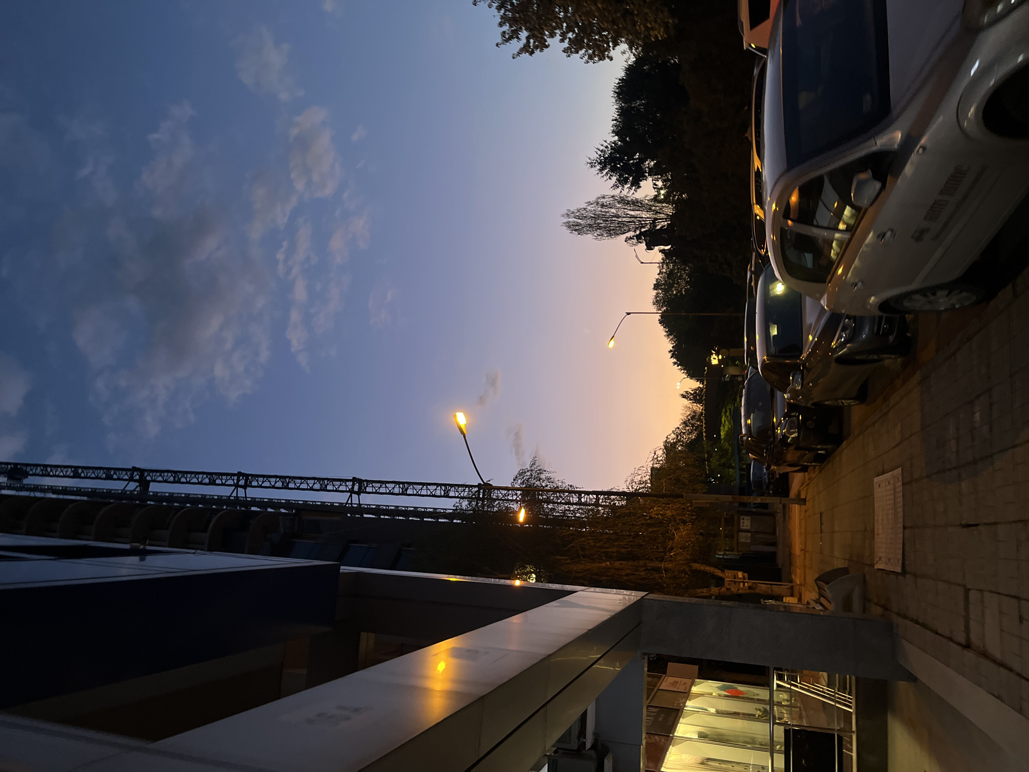 The image captures an urban street scene in Sofia, Bulgaria, at twilight. The sky dominates the upper portion of the frame, displaying a dramatic gradient from a deep indigo blue with scattered clouds on the left, transitioning to warm orange and pink hues towards the right horizon.
On the left, a modern building with dark facades and reflective surfaces occupies the foreground. Above it, a large, dark metal structure, possibly a bridge or part of a construction crane, is silhouetted against the sky. Several streetlights are illuminated throughout the scene, emitting a warm yellow glow and casting reflections on the building's surfaces and parked cars.
The right side of the image features a paved sidewalk running parallel to a row of parked vehicles. The closest car is silver, and the text "SAFETY MOTORS" is partially visible on its side. Silhouetted trees, some with sparse branches, line the street behind the cars. A white rectangular object, possibly a mat or tarp, lies on the sidewalk further down.
The scene conveys a sense of calm and quietude as day transitions into night. There is no visible human activity, presenting a static urban landscape illuminated by the natural glow of dusk and artificial streetlights.