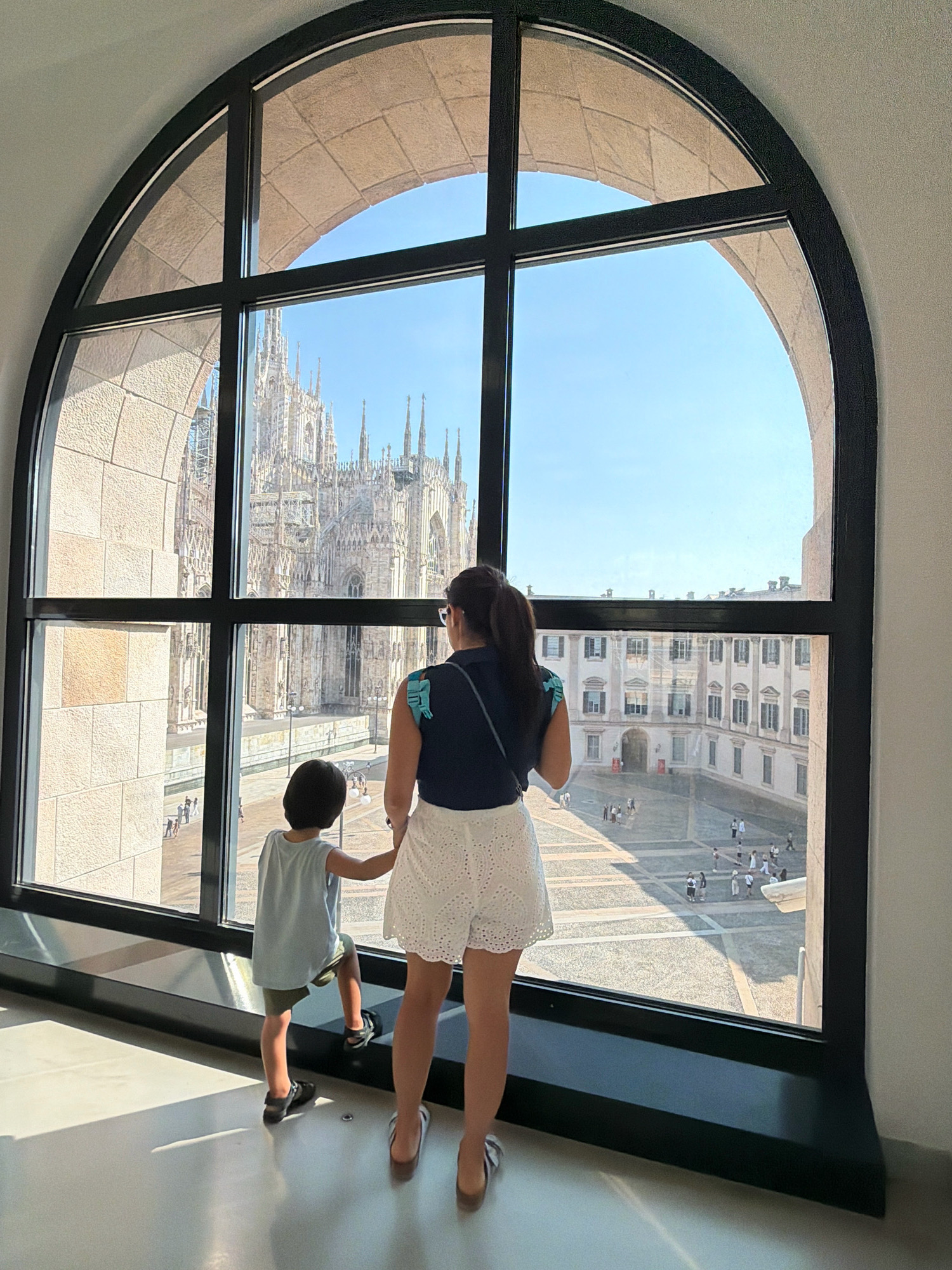 A woman and a child stand before a large, arched window, looking out at the Duomo di Milano (Milan Cathedral) in Milan, Italy. The woman, from behind, wears a dark top and white lace shorts, with her hair in a ponytail. The child, also facing away, is dressed in a light blue t-shirt and dark shorts. They are positioned on a ledge below the window, with sunlight casting shadows on the floor. The window offers a panoramic view of the Duomo and the surrounding Piazza del Duomo, with people visible in the square below. The architecture visible in the foreground, framing the window, suggests the view is from a building opposite the cathedral, possibly the Galleria Vittorio Emanuele II or a nearby palace.