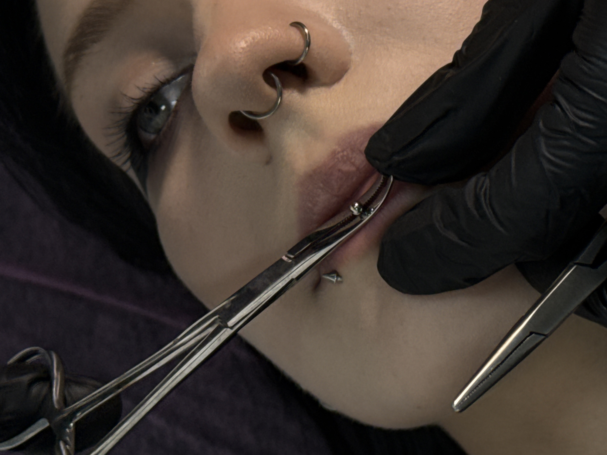 A close-up shot shows a person receiving a lip piercing. The person has a silver ring piercing in their nose. A silver labret stud is visible on the lower lip. The piercing is being performed by a person wearing black gloves, using a pair of silver piercing forceps to hold the lip in place. The background appears to be a dark purple fabric. The scene does not provide any indication of a location in Rostov-on-Don, Russia.