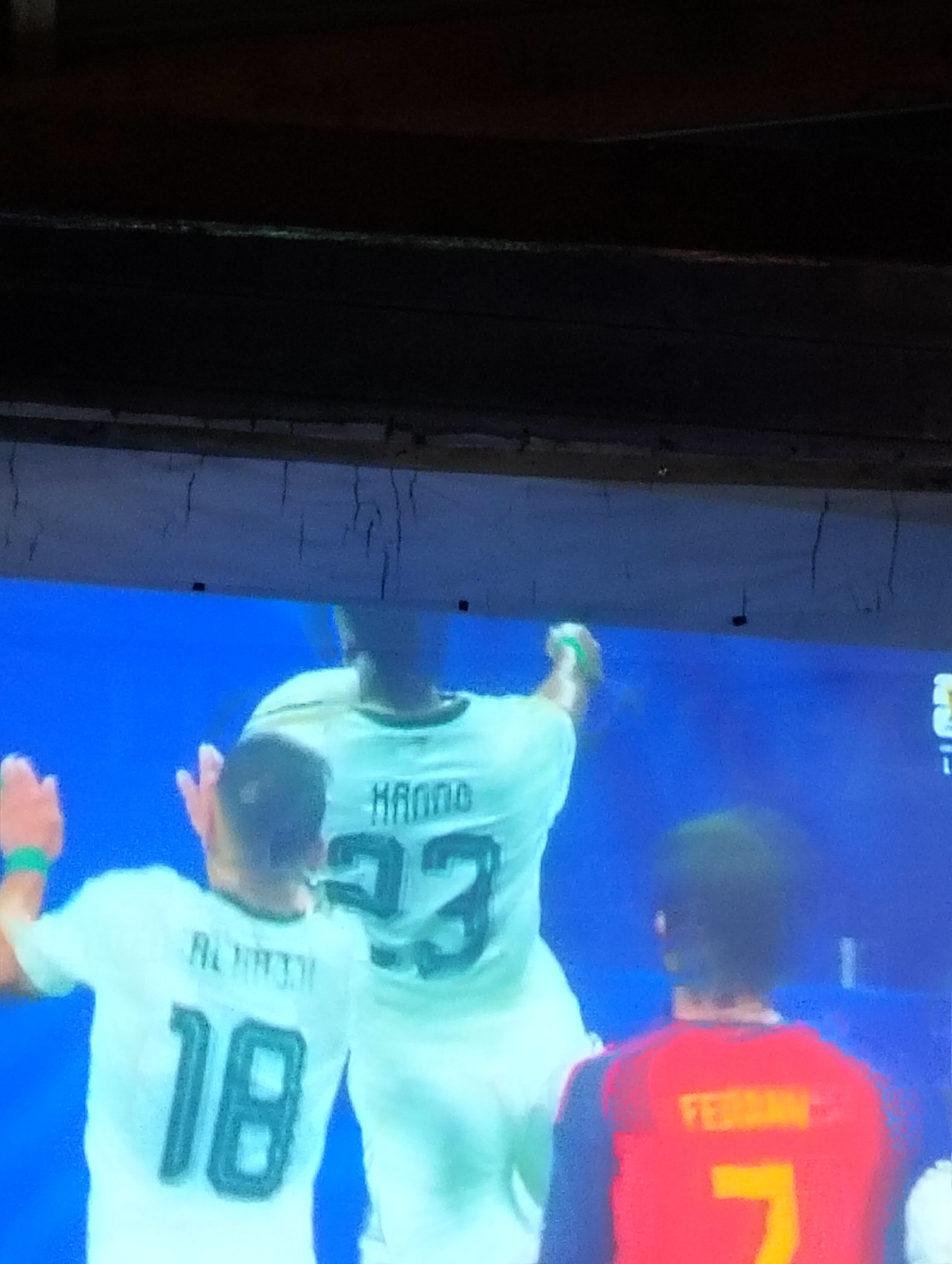 The image depicts a screen displaying a soccer match, with three players visible. The player in the foreground on the left wears a white jersey with the number 18. To his right is a player in a white jersey with "HANNO" and the number 33. On the far right, a player in a red jersey with "FERRAN" and the number 7 is partially visible. The background is a solid blue. The scene is viewed from below, with a dark, wooden structure partially obscuring the top of the image, suggesting it is being viewed on a television or projection screen in a dimly lit environment. No specific location in Payagaboh, Indonesia, can be identified from the image itself.