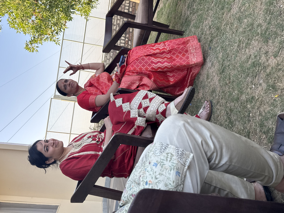 In Malasar, India, an outdoor scene depicts three individuals seated under clear skies. In the foreground on the left, a person, from the chest down, wears a light-colored kurta with a subtle floral pattern and shiny light-beige trousers, with dark brown sandals on their feet. To their right, a female, identified as mid-adult, is seated in a dark wooden chair, wearing a red kurta with gold and white embroidery and prominent silver-colored, triangular embellishments on her lower legs. She wears light-colored flip-flops and looks towards the viewer with a slight smile. To her right, an older adult female is seated in another dark wooden chair, dressed in a red sari with patterned gold and lighter red details. Her right hand is raised, fingers splayed in a gesture, and features mehndi; a dark mobile phone rests on her lap. She looks slightly to her left. The ground consists of green, somewhat sparse grass. In the background, a light-colored building wall with a window is visible to the left, and a metal grid fence extends across the middle and right, behind which green foliage and trees are present under a bright blue sky.